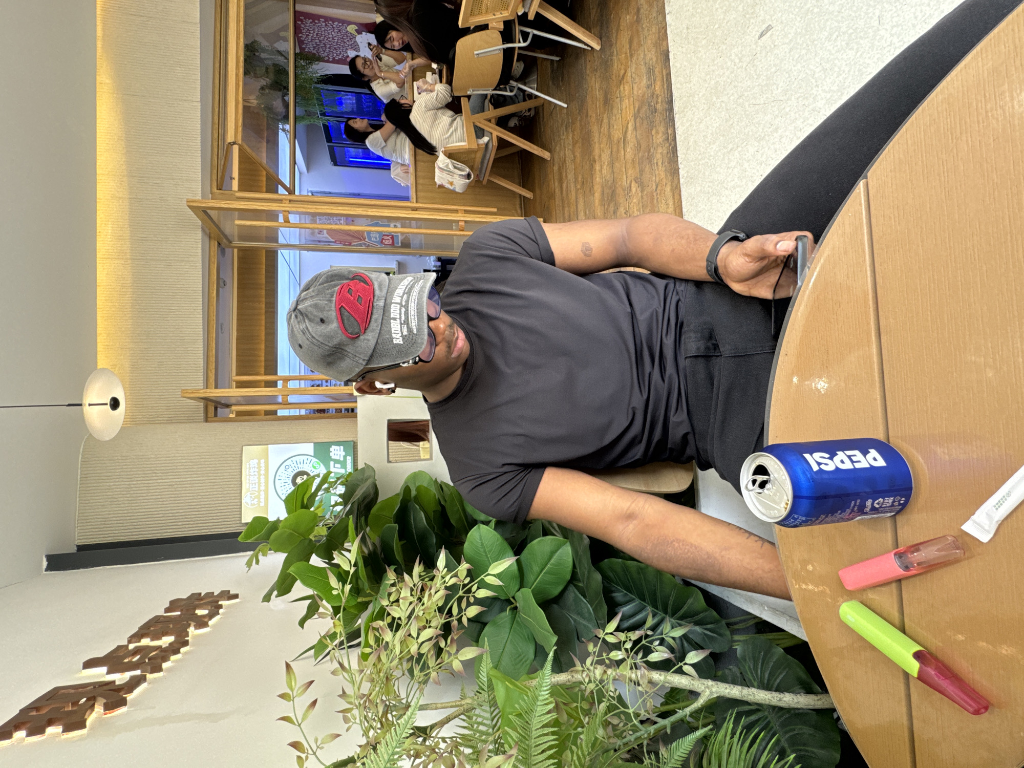 A medium shot captures a person in a black t-shirt and dark pants seated at a light brown table with greenery surrounding them, along with a can of Pepsi, a pink highlighter, a green highlighter, and a sugar packet. The person, wearing a gray cap, appears to be looking at a smartphone in their hand. The scene is likely in a restaurant in the Chenghua District of China, based on the signage visible in the background, which features Chinese characters. Other patrons can be seen in the background of the restaurant.