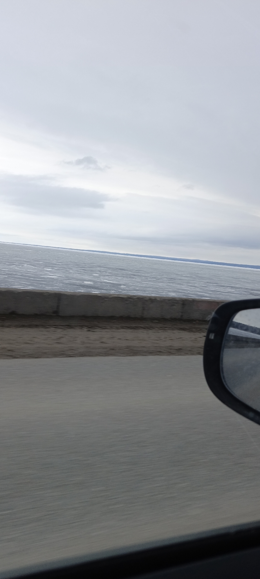 The image appears to be taken from inside a moving vehicle, showing a view of the Kama River in Chaikovsky, Russia. The river is mostly covered in ice, stretching to the horizon under an overcast sky. A concrete barrier separates the road from the riverbank. A side mirror of the vehicle is visible in the lower right corner of the frame, with reflections of the surrounding environment. The foreground shows the road surface, blurring due to the motion of the vehicle.