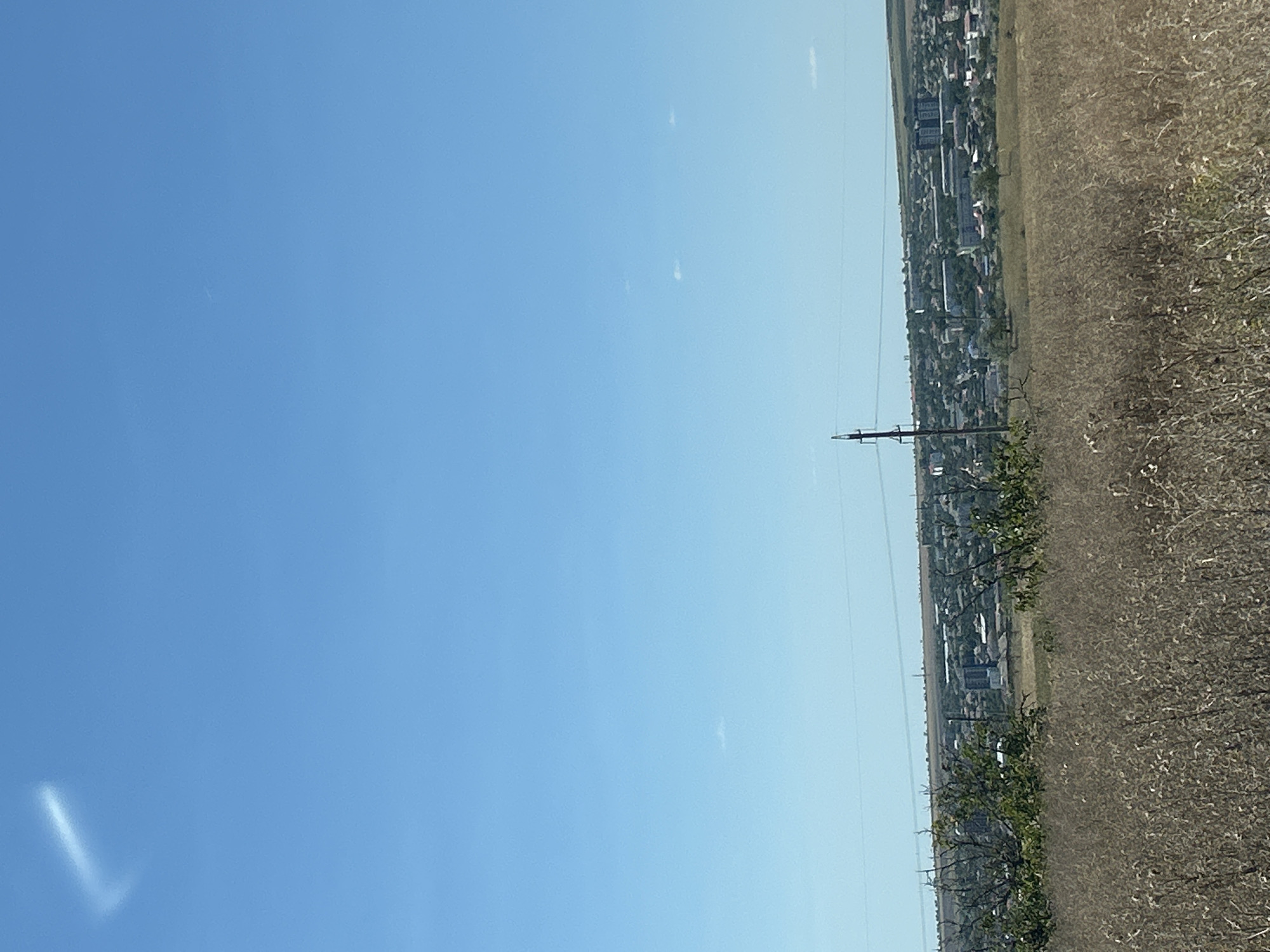 The image depicts a wide-angle view of a rural or semi-rural landscape with a town in the distance. The foreground is dominated by dry, golden-brown grasses and some sparse, bare-branched trees. In the mid-ground, a town can be seen nestled behind a line of trees and shrubs. The town appears to consist of a mix of low-rise residential buildings and some taller, modern apartment blocks. Power lines and a utility pole are visible in the mid-ground, indicating infrastructure. The background is a clear, bright blue sky with a few faint, wispy clouds. One prominent, elongated cloud formation, resembling a curved line or a hook, is present in the upper left quadrant of the sky. The overall impression is a sunny day in a relatively undeveloped area, possibly on the outskirts of a town like Comrat, Moldova, given the context. There are no people or animals visible in the image.