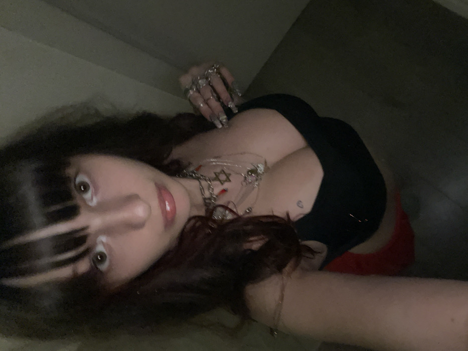 A woman with dark hair and blunt bangs is captured in a close-up, low-angle shot. She wears a black top, and red shorts are slightly visible. Her hand is adorned with multiple rings. Necklaces featuring a Star of David and other charms decorate her neck. A small heart tattoo is visible on her chest. She has fair skin, full lips with pink lipstick, and her eyes are wide. The lighting is dim, creating shadows on her face and background.