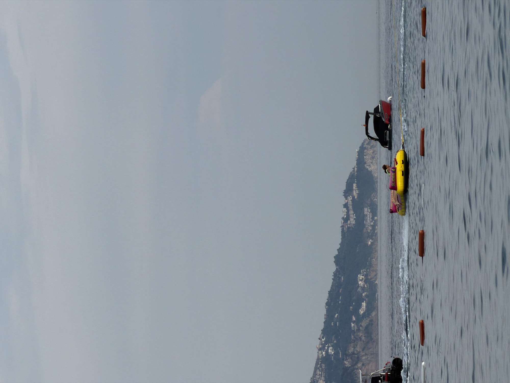 The image depicts a serene scene on the water, likely in Budva, Montenegro, with a mountainous coastline dotted with buildings in the background. The sky is overcast, suggesting a cloudy or hazy day.
The primary subjects are a yellow inflatable tube with a person on it, being pulled through the water, and a black and red speed boat. The person on the tube appears to be enjoying a recreational activity. The boat is positioned behind the tube, likely towing it. There are also several orange buoys floating in the water, marking a course or area.
The activity is water sports, specifically tubing, with a speed boat providing the propulsion. The overall mood is relaxed and leisurely, consistent with a day at the beach or on the water. The time of day is not explicitly clear due to the overcast conditions, but it appears to be daytime. The location cues, like the distinct mountainous coastline with white buildings, are characteristic of the Adriatic coast, and specifically Budva.
There is some visible text on the yellow inflatable tube, which reads "SUPER 4".