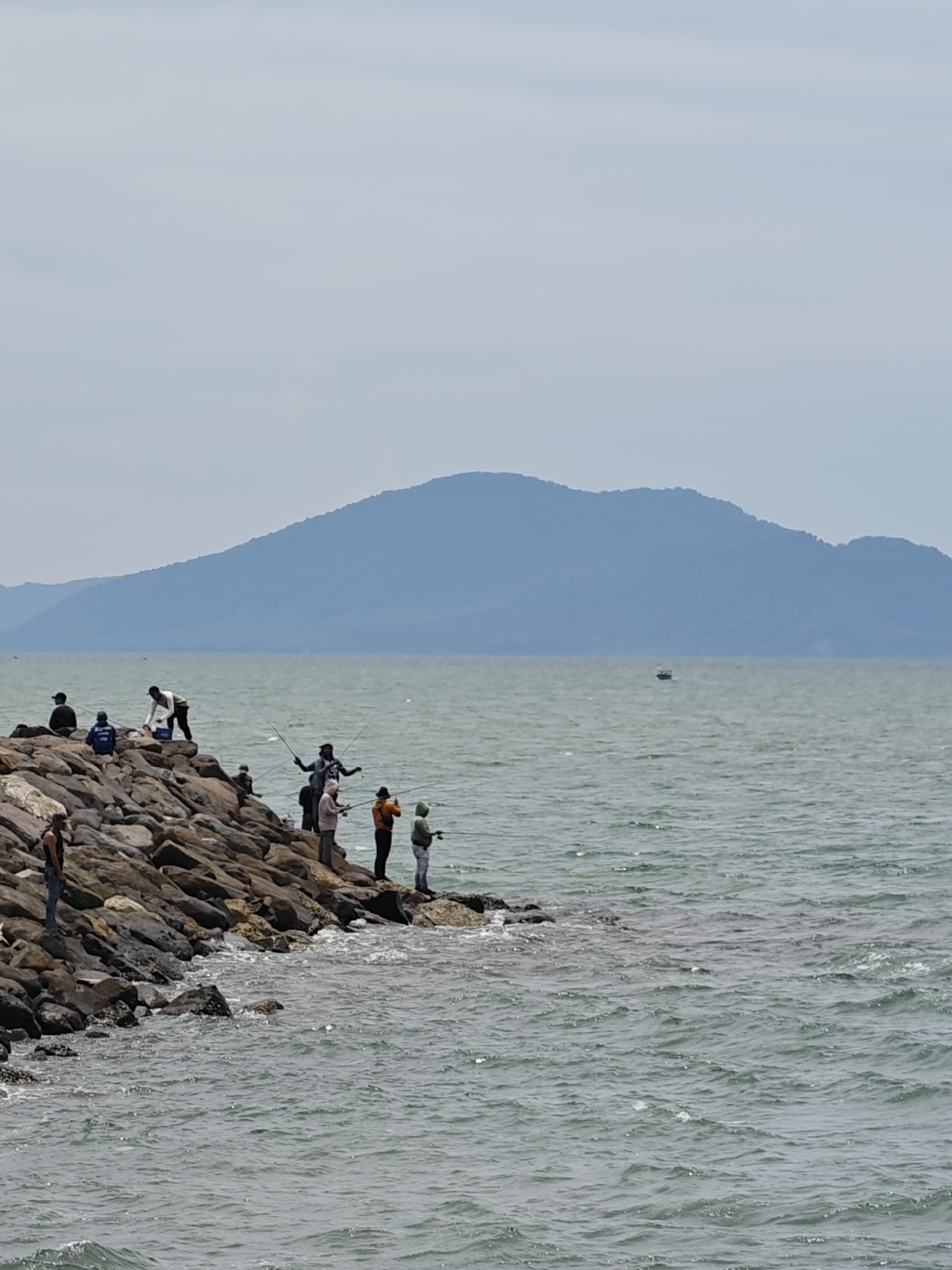 The image depicts a group of people fishing on a rocky breakwater in Banda Aceh, Indonesia. A mountain can be seen in the background, across the ocean. The sky is overcast. Several people are positioned along the breakwater, holding fishing rods, actively engaged in fishing. Some individuals are standing, while others are seated. The breakwater is constructed of large rocks, and waves are lapping against it. A small boat is visible on the water in the distance.
