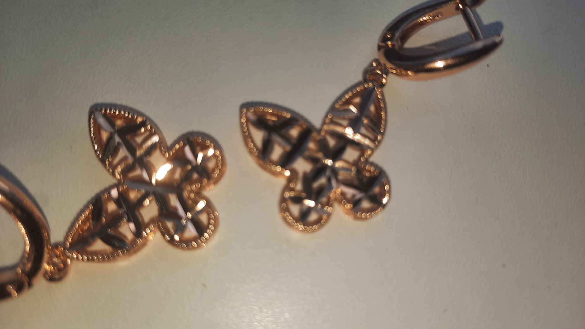 The image displays a close-up, overhead view of a pair of rose gold earrings with butterfly-shaped pendants. The earrings are on a light-colored, textured surface, possibly a table or countertop. The lighting creates reflections and highlights on the metallic surface of the jewelry, emphasizing its intricate, cut-out design. One earring is positioned to the left, with its pendant slightly to the left of the frame. The other earring is to the right, with its pendant to the right of the frame and its clasp visible at the top right. There are no people or any contextual elements to suggest a location in Shklov, Belarus, or any other specific geographic setting. The focus is solely on the jewelry.