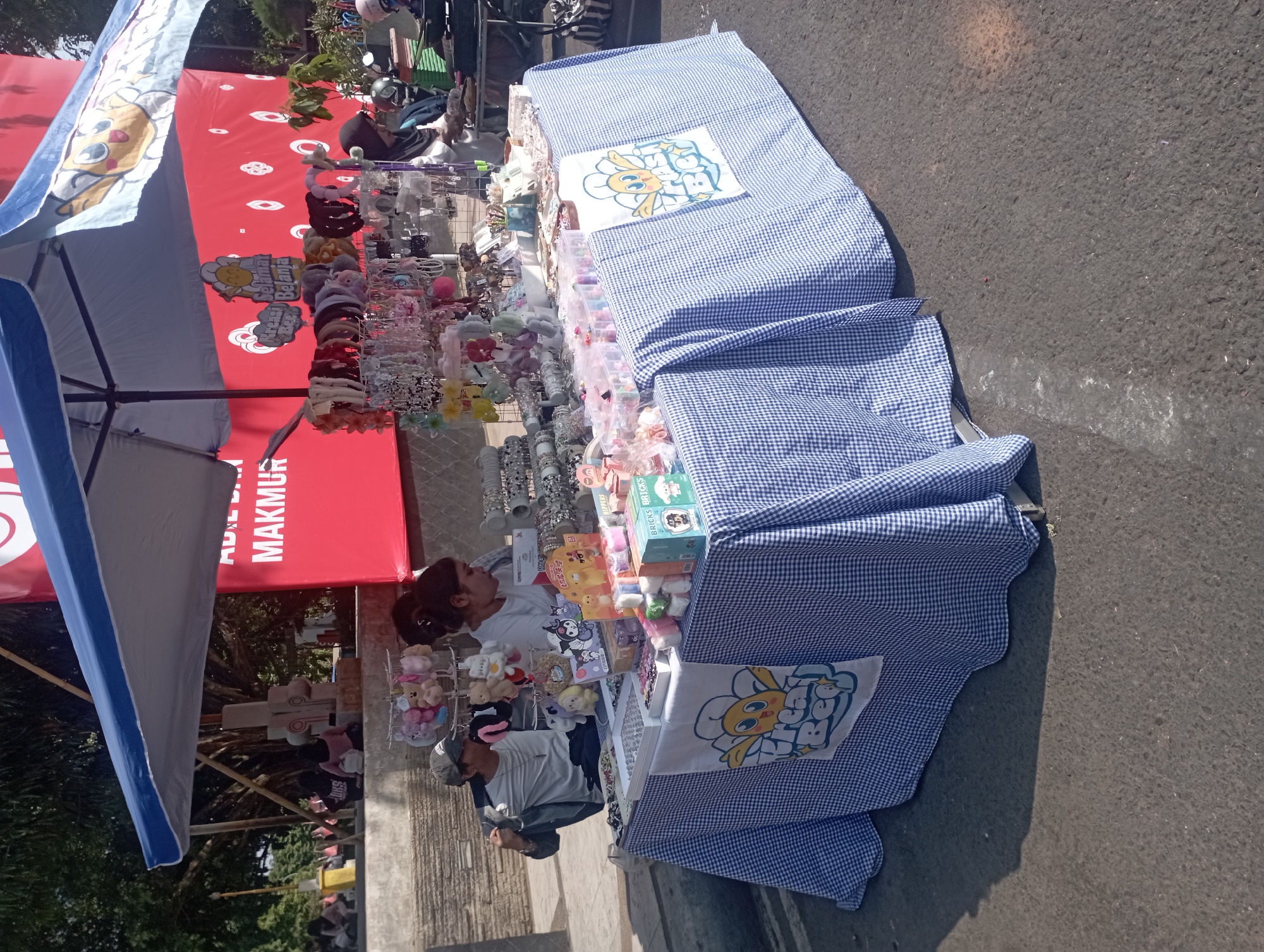 The image shows an outdoor market stall in Nganjuk, Indonesia. The stall is set up with a blue and white checkered tablecloth and is covered by a blue and white awning. A variety of items are displayed on the table, including hair accessories, small plush toys, and what appear to be stationery items. There are two people visible at the stall; one person, wearing a grey t-shirt and a baseball cap, is sitting and looking towards the right side of the image, while another person, wearing a white t-shirt and a headscarf, is standing and appears to be attending to the stall. In the background, there is a red banner with white text and graphics, and some trees and other market activity can be seen. The stall is located on an asphalt surface, suggesting it is on a street or public area within the city.