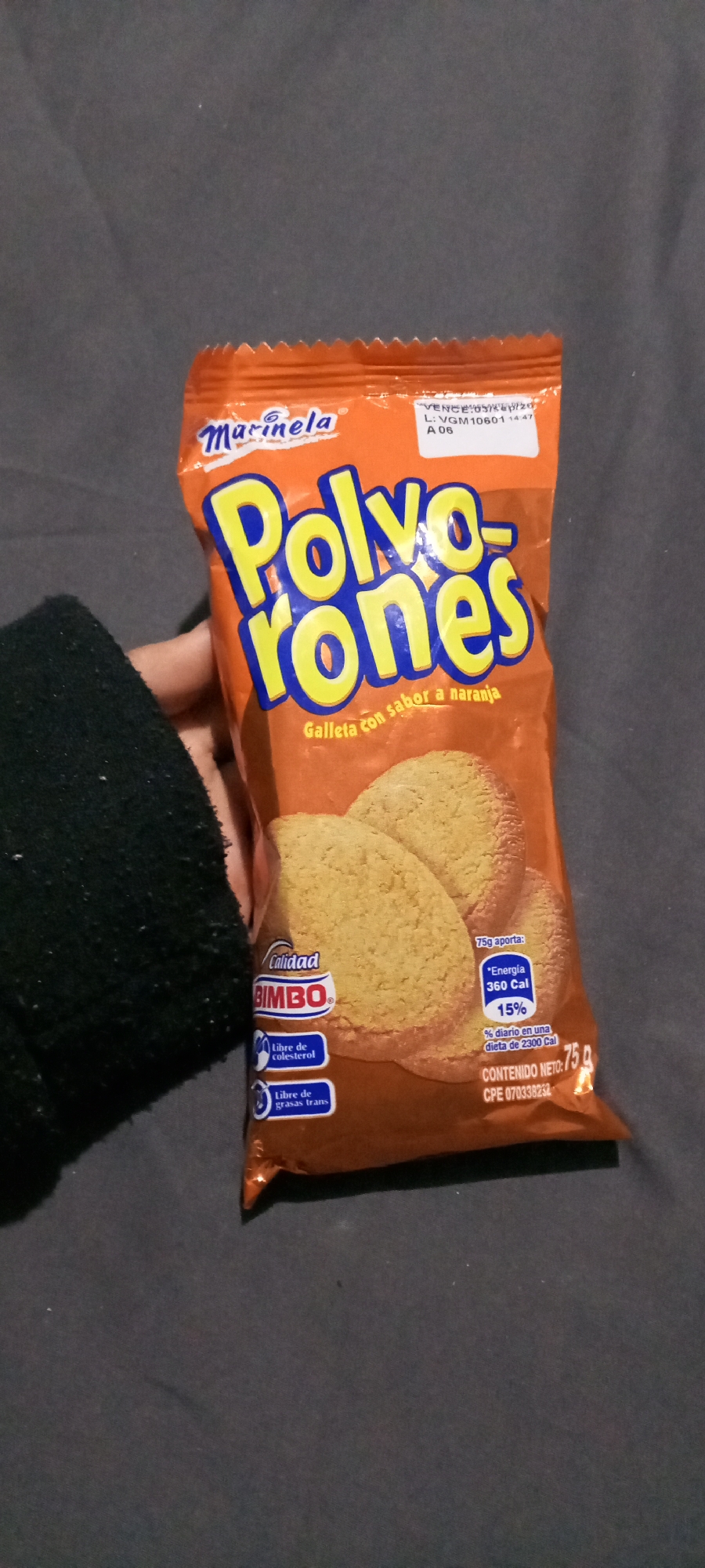 A package of Marinela Polvorones cookies is being held in someone's hand. The packaging is orange and features the brand name Marinela at the top, along with the product name "Polvorones" in large, blue and yellow lettering. Below the name, it says "Galleta con sabor a naranja," indicating orange-flavored cookies. The front of the package displays images of the cookies. The package also has details like "Calidad Bimbo," "Libre de colesterol," and "Libre de grasas trans." Nutritional information, including the caloric content and percentage of daily intake, is provided. The net content is listed as 75 grams. The expiration date is visible as "Vence: 05/sep/20," and the lot number is "L: VGM10601 14:47 A 06." The background is a dark gray, possibly a fabric surface. The person holding the package is wearing a dark-colored sweater or sleeve.
The information provided does not mention the location of Juan Griego, Venezuela.