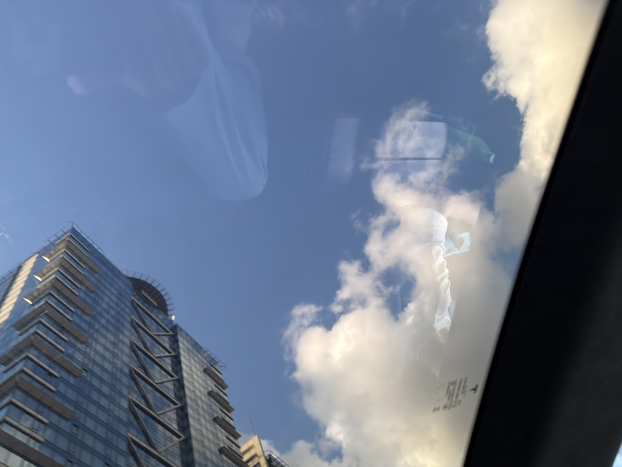 The image presents an upward view of a modern urban landscape and sky, captured through a reflective glass surface, likely the sunroof or windshield of a vehicle. On the left, a tall, contemporary building with a blue glass facade rises into the frame. It features multiple balconies and a helipad on its roof, along with prominent diagonal architectural elements.
The background shows a clear blue sky dotted with scattered white, sunlit clouds, indicating a bright, sunny daytime.
The glass surface exhibits significant reflections. In the upper-left area, the faint silhouette of a person's head and shoulder is reflected. Towards the center and right, more distinct reflections include a white, fabric-like object, possibly a mask, and a vibrant green object. In the bottom-right corner of the image, a small rectangular label is reflected, clearly showing a "T" logo and the word "TESLA," confirming the viewpoint is from inside a Tesla vehicle. The scene depicts an outdoor setting in Sofia, Bulgaria.