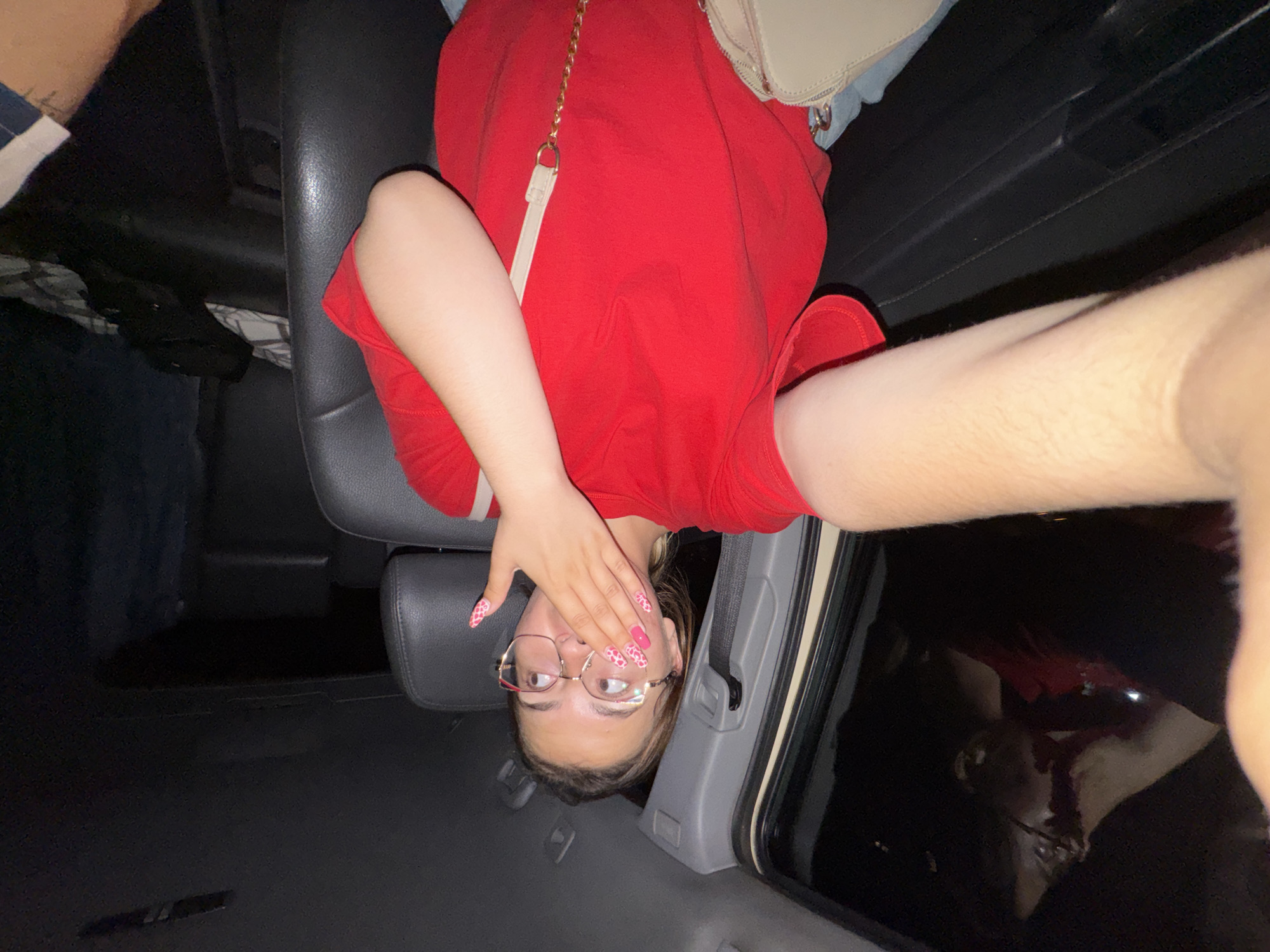 A young woman, seated in the passenger seat of a vehicle, holds her left hand over her mouth, her eyes wide with surprise or shock as she looks to her left, out of frame. She is wearing a bright red t-shirt, blue denim shorts, and distinctive eyeglasses with large, pink-rimmed frames. Her nails are long and painted with a pink and white pattern. A light-colored purse with a gold chain strap is visible in the lower part of the frame. The interior of the car, upholstered in dark material, is dimly lit, with the outside environment appearing dark, suggesting it is nighttime. The woman's right arm is extended towards the camera, likely holding a phone to take a selfie.