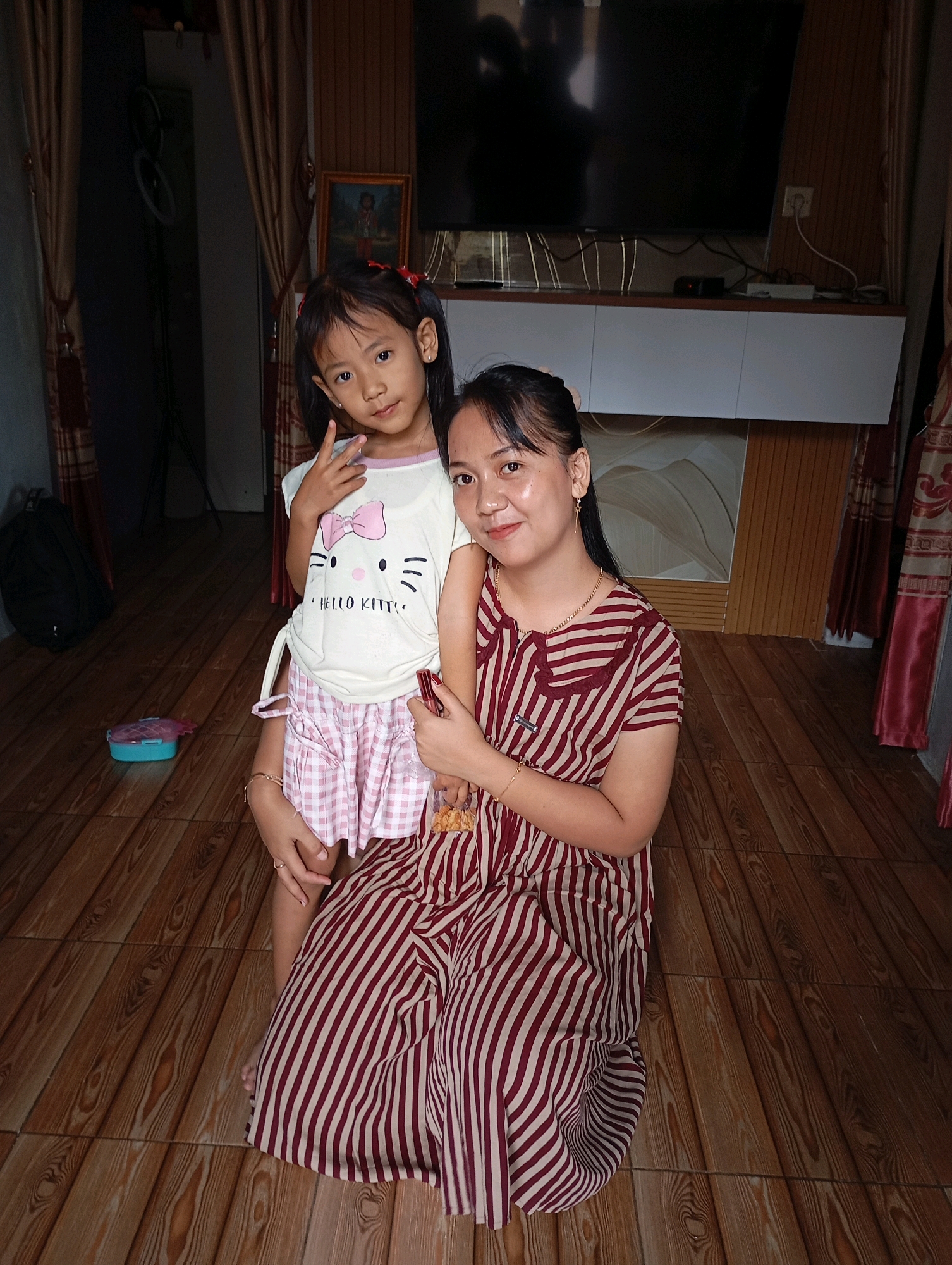 In an indoor setting, likely a living room, a woman kneels on a wooden floor next to a young girl. The woman is wearing a red and white striped dress, and the girl is wearing a white shirt with a pink Hello Kitty graphic and pink and white checkered shorts. The girl is holding up two fingers in a peace sign. The room has wood-paneled walls, a television mounted on the wall, and curtains. There is a black backpack on the floor to the left, and a small pink container. The scene appears to be a casual, everyday moment captured in a residential interior.