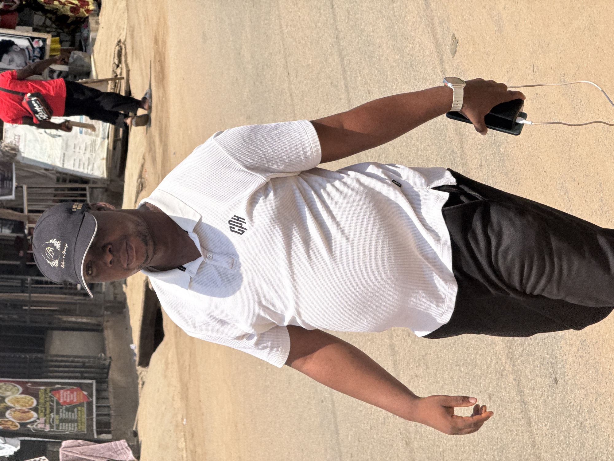 A man, wearing a white polo shirt and a dark baseball cap, stands on a paved surface in a sunlit outdoor setting. He is holding a dark smartphone and a white cable, which appears to be connected to a power bank. The man is looking towards the camera. In the background, a person in a red shirt and black pants is partially visible, walking away from the camera. To the left of the frame, a stall with various signs and images, possibly for food, is visible. The scene suggests a daytime setting, likely in a market or busy street. The lighting indicates it is a bright day. The clothing of the man and the overall environment suggest a warm climate. The text "GBK" is visible on the man's shirt.