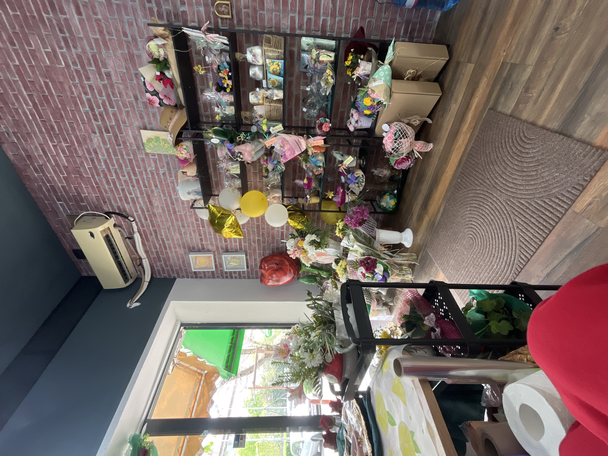 The image shows the interior of a flower shop with shelves stocked with bouquets, arrangements, and gift items against a brick wall. Several balloons are visible, including yellow and gold colors. The shop is filled with plants, flowers, and wrapping materials, suggesting a busy, creative workspace. A window lets in natural light. Based on the provided context, this image appears to be a store in Burgas, Bulgaria.