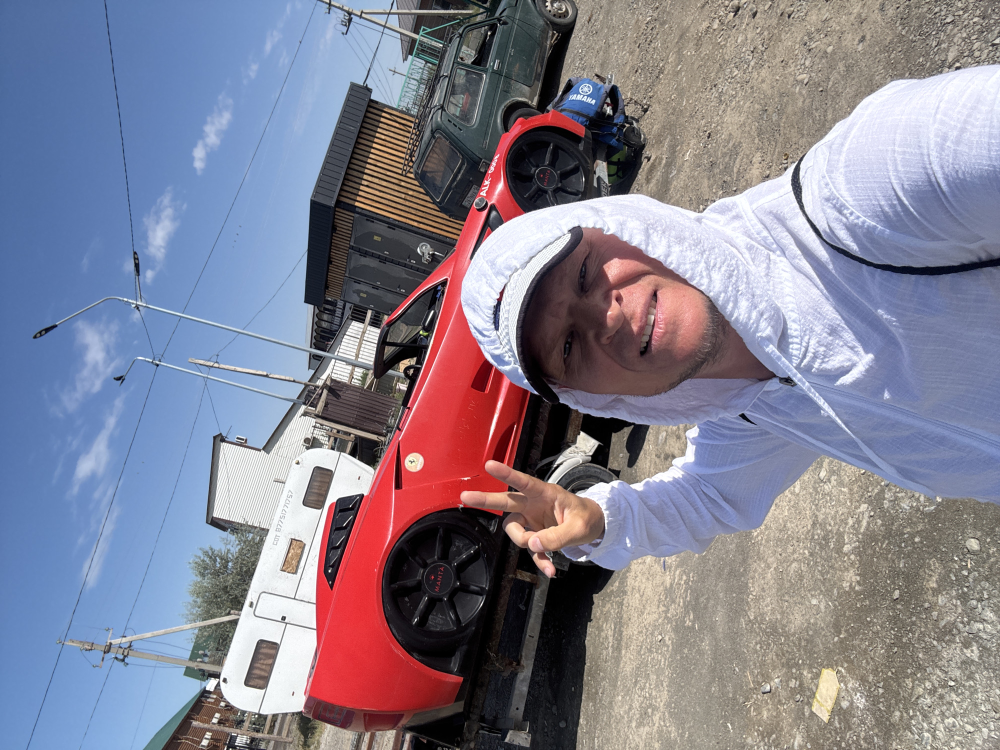 A man in a white hooded shirt takes a selfie in front of a red car with a "MANTA" logo on its wheel. The car is on a trailer, next to a white camper and a green vehicle. The scene appears to be in an outdoor, somewhat industrial area. The background shows buildings, power lines, and a blue sky with clouds. The man is making a peace sign with his right hand. The car seems to be a replica or kit car. The text on the camper, "COT. 87751771757," is partially visible. The location is identified as Zharbulak, Kazakhstan.