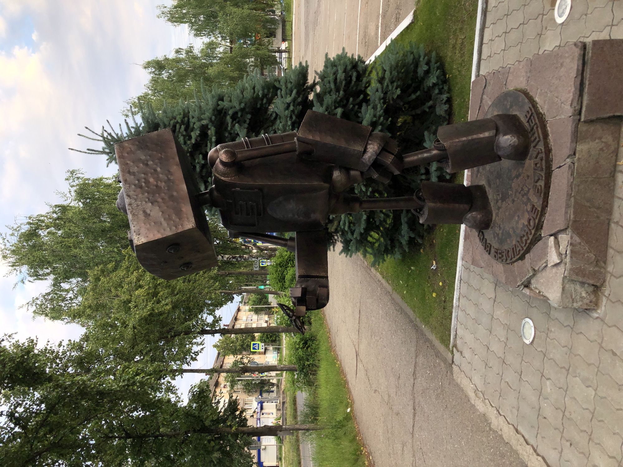 The image shows a bronze statue of a robot in the city of Chaikovsky, Russia. The robot is standing on a circular metal base inscribed with Russian text. The robot is made of various metallic components, giving it a rugged and industrial appearance. It has a large, box-shaped head, a torso with mechanical detailing, and sturdy limbs. The robot is positioned on a paved area surrounded by greenery, including trees and bushes. In the background, there are multi-story buildings, a street with a pedestrian crossing, and a parking lot. The sky is visible above, with clouds. The overall setting suggests an urban environment, likely a public space within the city.