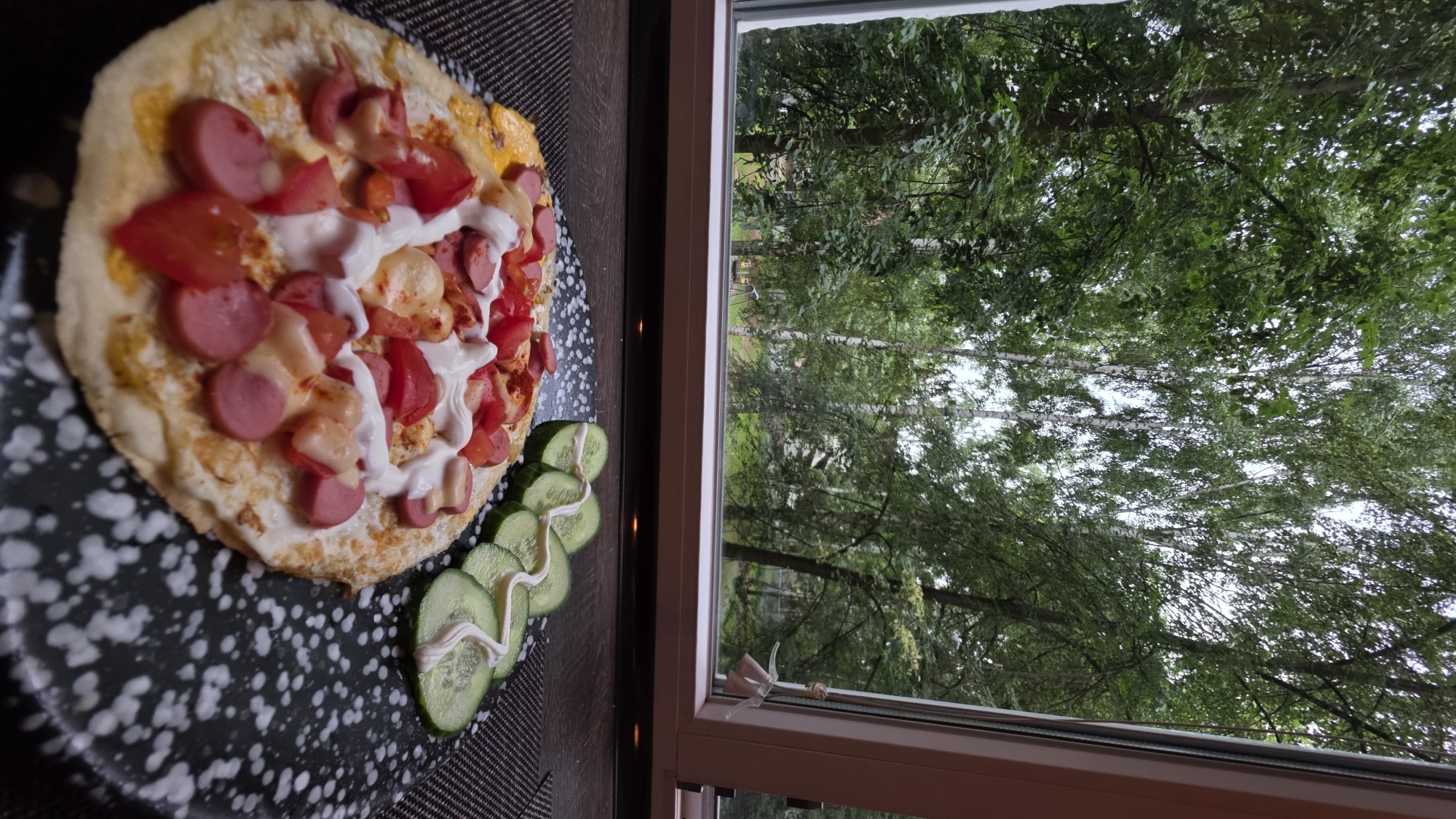 A pizza with sliced sausages, tomatoes, and white sauce is placed on a dark, speckled plate next to sliced cucumbers drizzled with sauce. The plate sits on a dark placemat. To the right of the pizza is a window offering a view of a lush green forest. The trees are dense with leaves, and some birch trees with white bark are visible. The overall lighting suggests it is daytime, and the abundance of greenery implies a warm or temperate climate, possibly summer or late spring. The scene appears to be set indoors, looking out at a natural landscape. There is no visible text in the image.