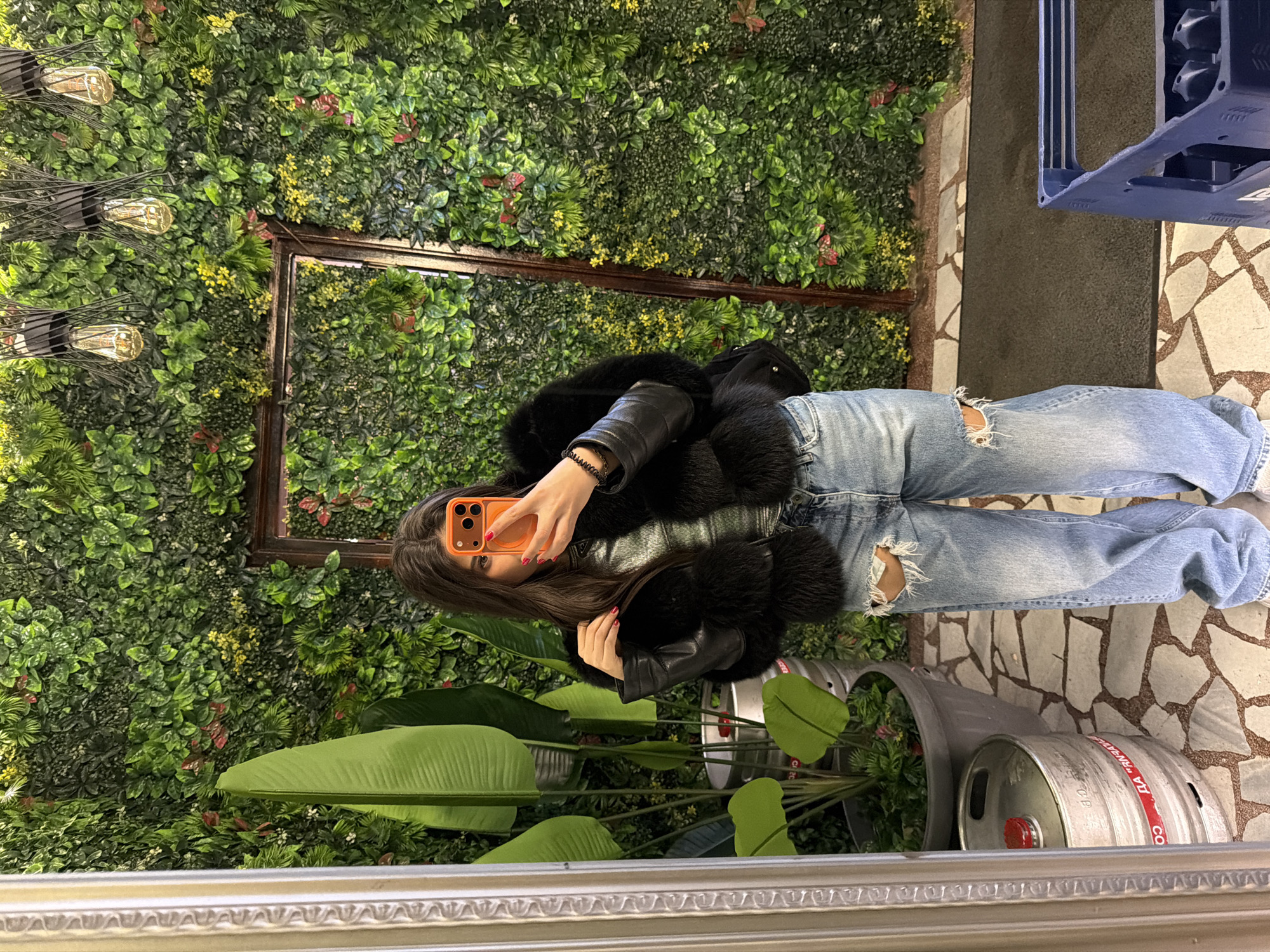 A young woman takes a selfie in front of a wall decorated with greenery and flowers. She wears a black fur-trimmed jacket and ripped blue jeans. The photo is taken indoors, with a mirror framing the scene. The location appears to be in Rudartsi, Bulgaria, based on the presence of a keg bearing Cyrillic text, commonly used for alcoholic beverages in Bulgaria. A large potted plant with broad leaves is visible in the foreground, and a decorative metal frame is on the wall behind the woman. The floor has a mosaic tile pattern.