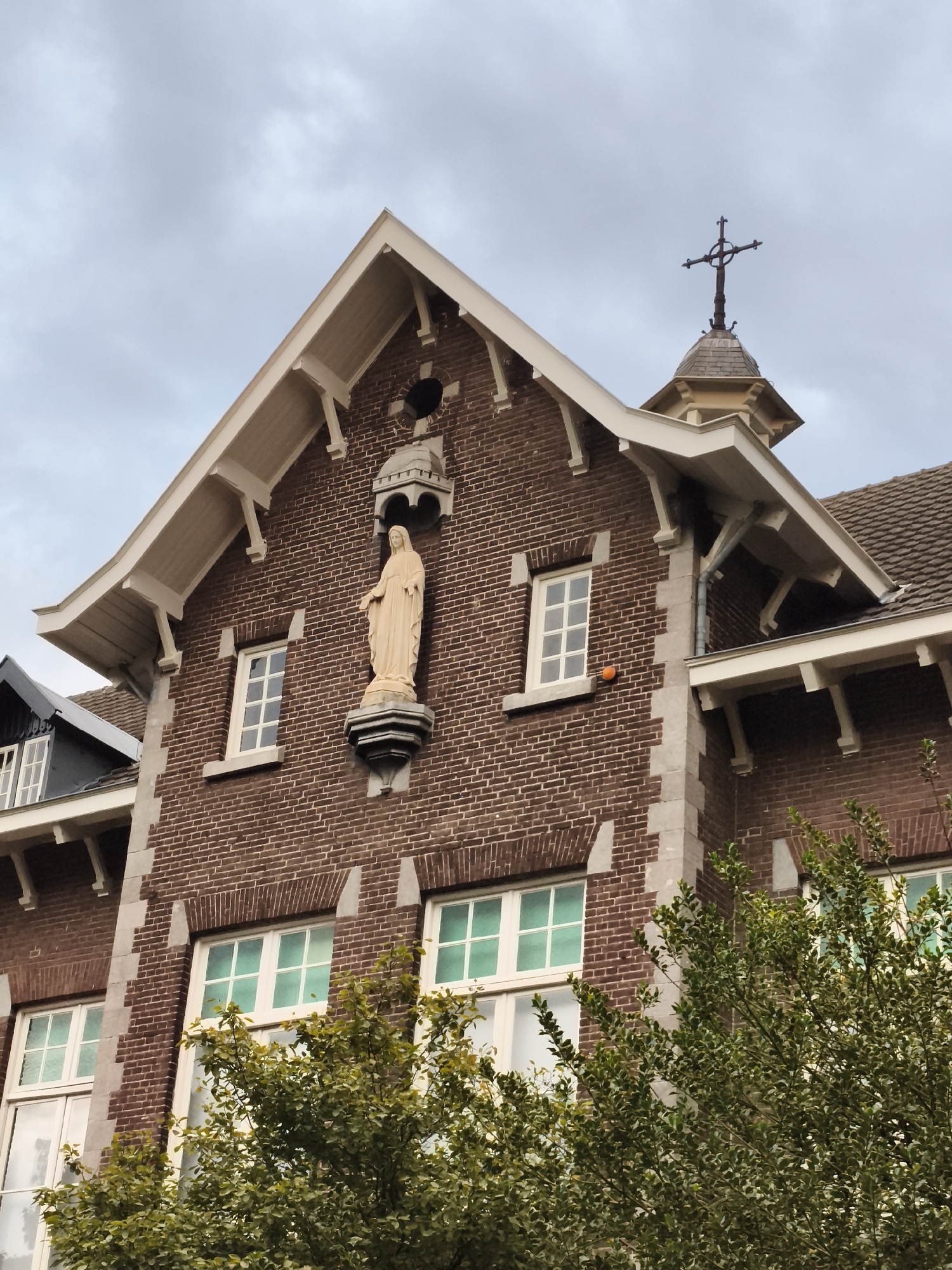 The image shows a close-up, low-angle view of the facade of a brick building with architectural details suggesting it is a religious institution, possibly a former church or school, in Maastricht, Netherlands. The building features a prominent statue of the Virgin Mary positioned on a stone pedestal, centrally located on the upper portion of the brick wall. Above the statue, a small, circular opening is visible within the brickwork. The roofline is accentuated by white eaves and decorative brackets. A cross is mounted on a small, octagonal turret atop the building. Several windows with white frames and greenish panes are visible on different levels. Lush green trees with dense foliage partially obscure the lower portion of the building. The sky is overcast with gray clouds. The overall impression is of an old, possibly historic, religious building.