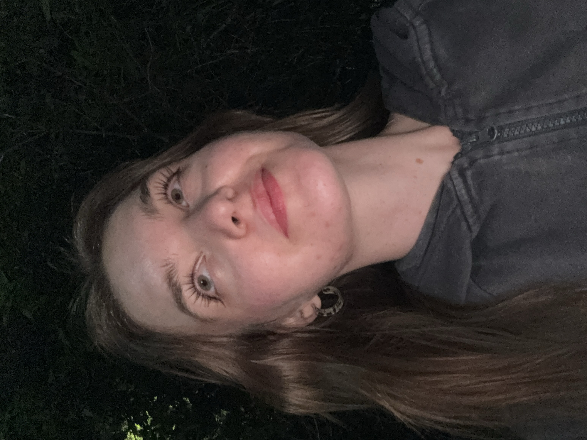 A woman with light skin, light brown hair, and light eyes poses for a selfie-style photo outdoors at night. She is wearing dark lipstick and a grey zipped jacket. Her hair is long and wavy. She has an earring in her left ear. The background appears to be dark, possibly foliage. The location in Perm, Russia is not discernible from the image.