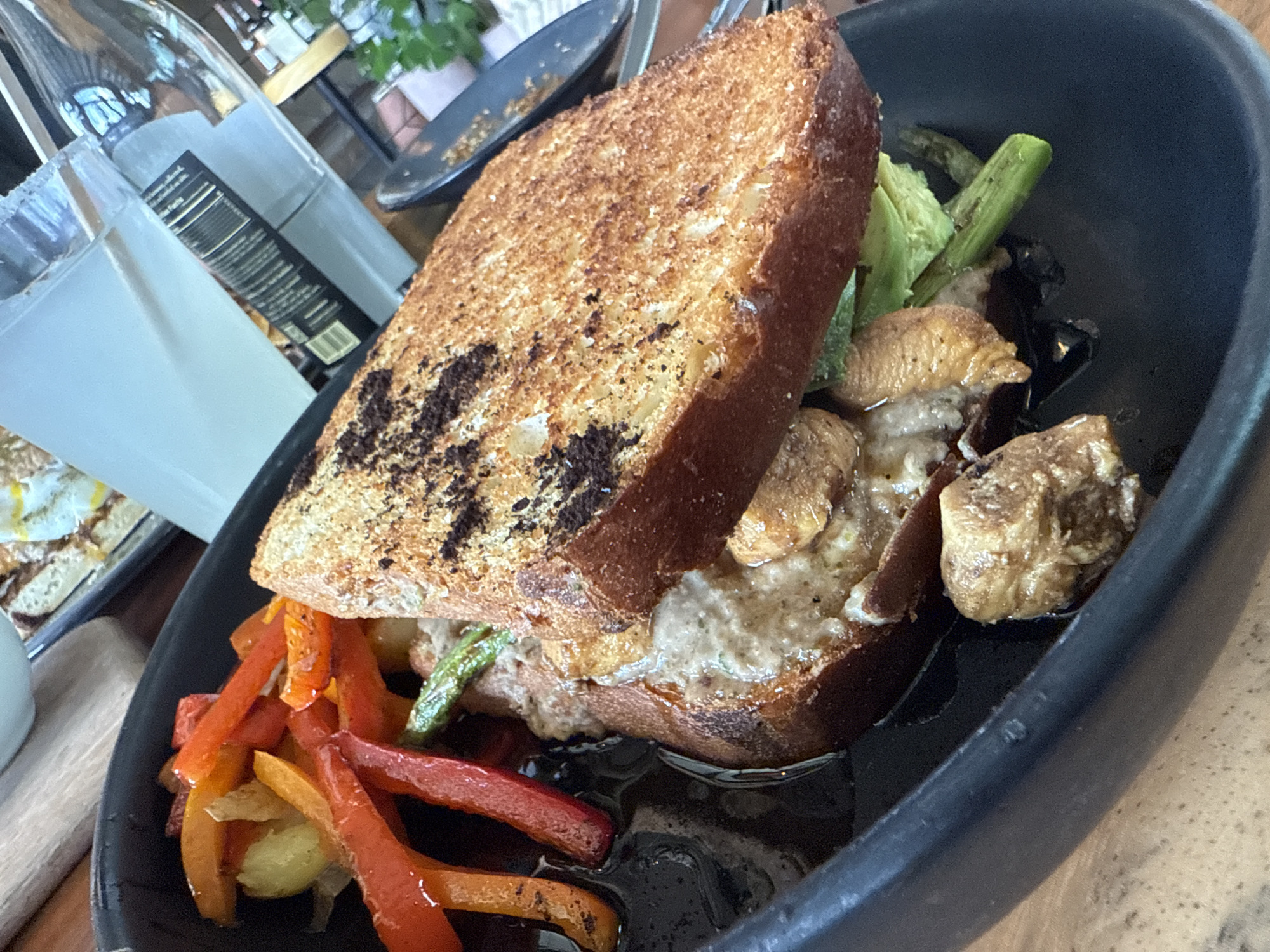 A close-up shot of a plate of food, most likely taken in San Andrés Cholula, Mexico, given the location in the prompt. The main feature is a thick sandwich with heavily toasted bread, served in a black bowl. The sandwich contains visible ingredients such as asparagus, a creamy spread, and sautéed mushrooms or a similar protein. Beside the sandwich are strips of sautéed bell peppers in red and orange hues. The food seems to be sitting in a liquid, possibly a sauce. In the background, a glass bottle of a clear liquid is partially visible, alongside another plate of food slightly out of focus. The angle of the shot suggests that the photograph was taken from above. The lighting is natural, highlighting the textures and colors of the dish.