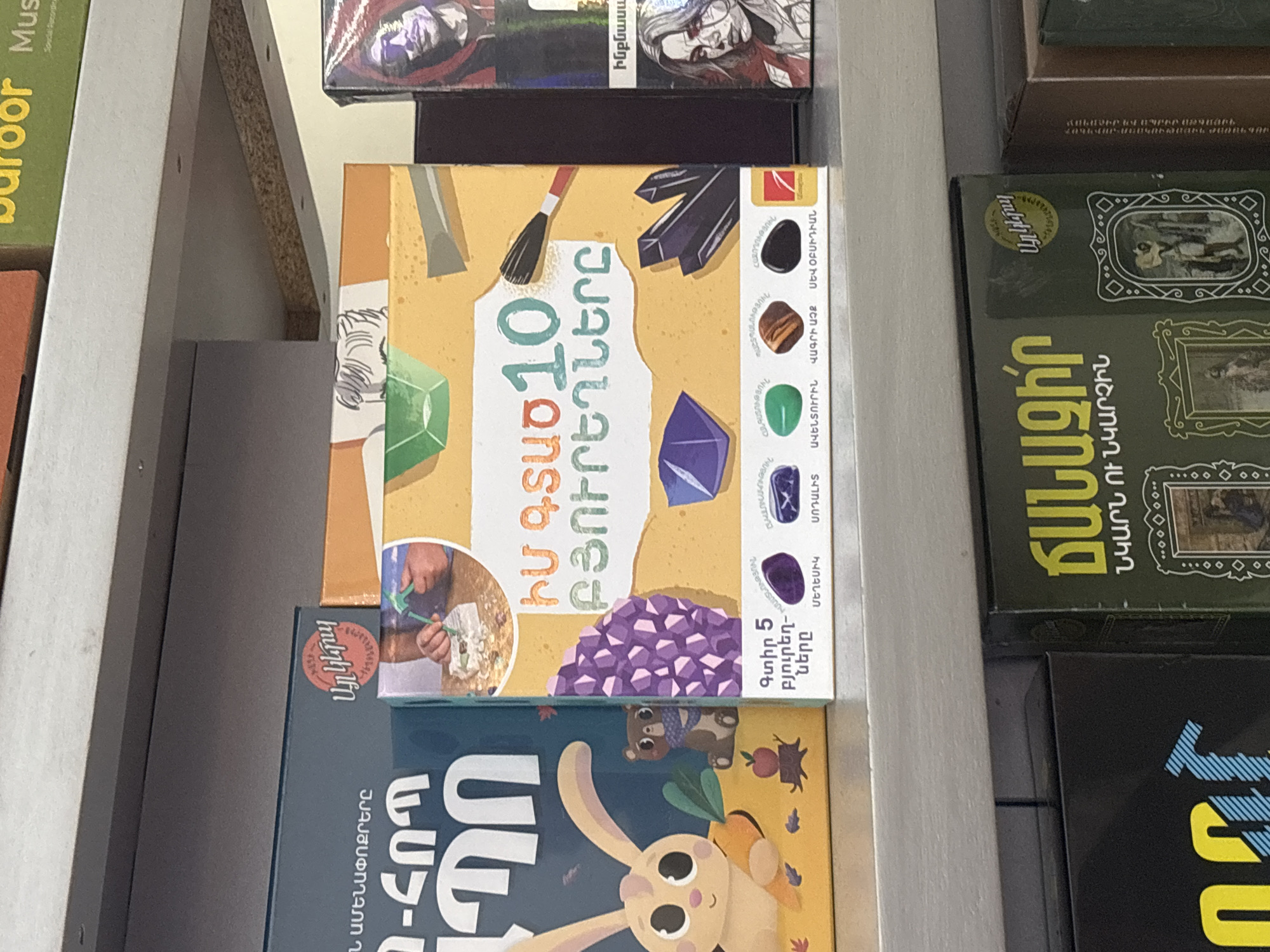 This image captures a detail of shelves filled with various boxes, likely board games or educational kits, in Yerevan, Armenia. The scene is indoors, brightly lit, focusing on a horizontal stack of items.
The most prominent item is a rectangular box for a children's activity kit, oriented vertically on the shelf. Its design features illustrations of tools like a pickaxe and brush, alongside various crystals in green, purple, and black hues. A child's hands are depicted digging in sand with a small shovel, suggesting a "digging for crystals" activity. Above a large cluster of purple crystals, text in Armenian reads "ԻՄ ԳՏԱԾ 10 ԲՅՈՒՐԵՂՆԵՐԸ" (My Found 10 Crystals) and "Գտիր 5 բյուրեղները" (Find 5 crystals). A list of crystals including Malachite, Aventurine, Tiger's Eye, Black Obsidian, Sodalite, Amethyst, and Morion (a type of smoky quartz) is also visible. The brand logo "Այլ կերպ" (Ayl Kerp) is present.
Below this main box is another, partially visible box featuring cartoon characters, a rabbit and a bear, with text "ԱՄԵՆԱՓՈՔՐԵՐԸ" (The Smallest Ones) and "ՆՄՈՒՇ" (Sample/Model), indicating a children's game or book for younger audiences.
To the right, a dark green box displays framed pictures and the title "ՃԱՆԱՉԻՐ ՆԿԱՐՆ ՈՒ ՆԿԱՐՉԻՆ" (Recognize the Painting and the Painter), also by "Այլ կերպ" (Ayl Kerp). Above the main box, other boxes are stacked, one showing a stylized human face and partial Armenian text "Ինքնաս" (Self-...), possibly from a game title. English text fragments "Mus" and "Recording" are also partially visible on other boxes, suggesting a diverse collection of media.