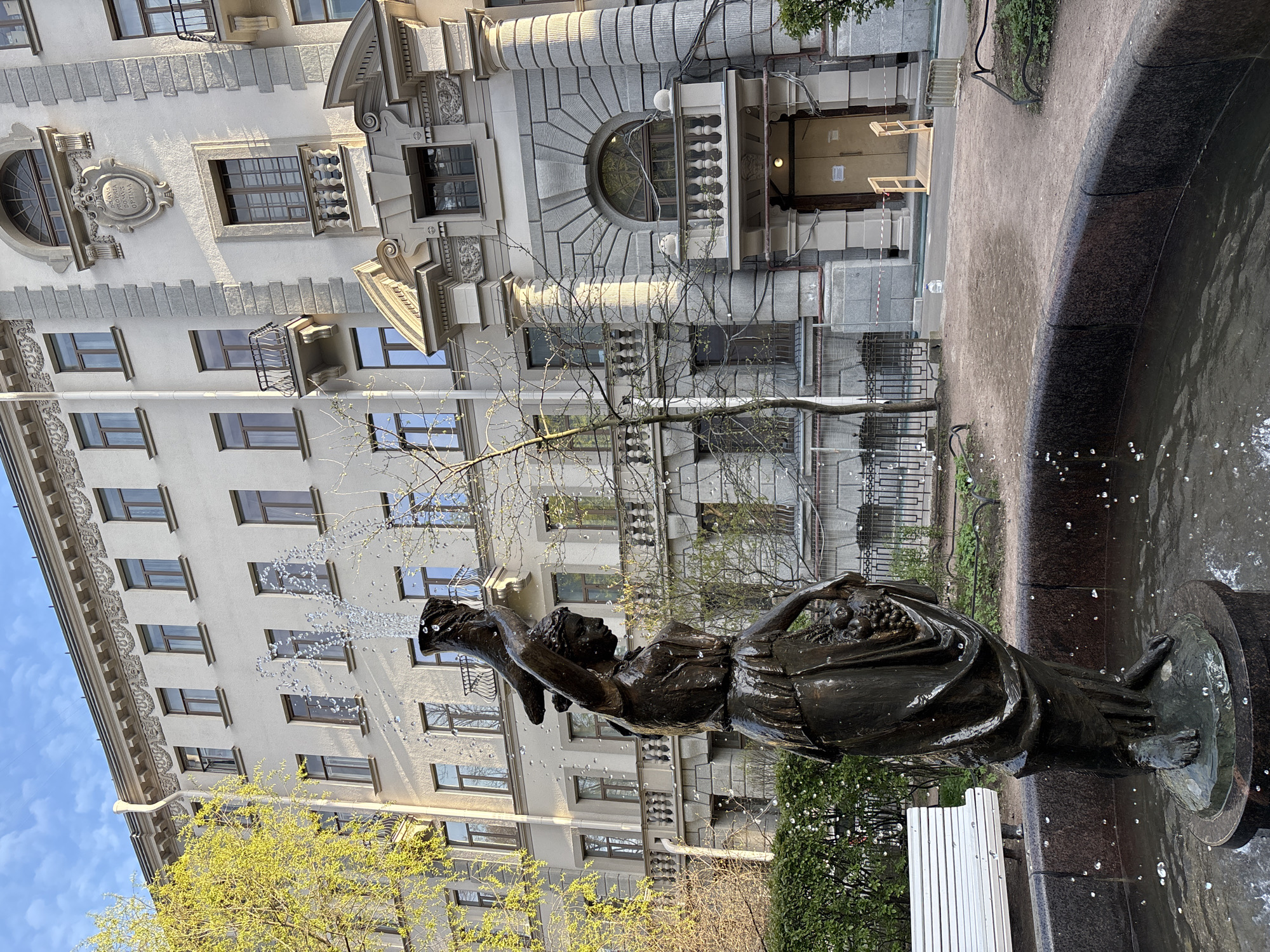 The image shows a bronze statue of a woman located in a fountain in Saint Petersburg, Russia. The statue is positioned in front of a multi-story building with architectural details such as balconies, decorative windows, and ornate stonework. Water is spouting from the fountain near the statue's head, creating a spray in the air. In the foreground, parts of the fountain basin and edges are visible. To the right of the statue, there's a white bench. The surrounding environment includes trees and vegetation, suggesting the fountain is in a park or garden.