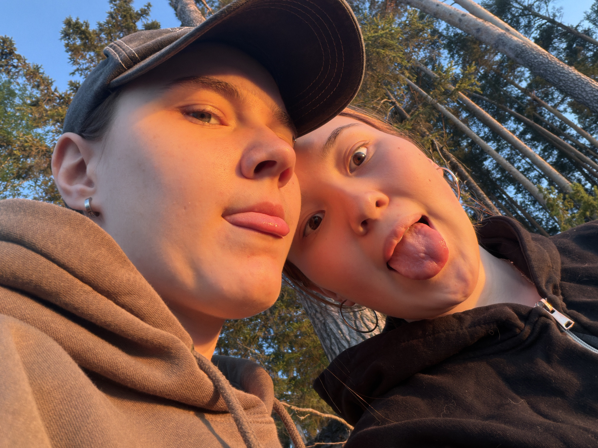 Two individuals are in the foreground of a selfie taken outdoors. The person on the left wears a gray baseball cap and a gray hoodie. Their tongue is sticking out slightly. The person on the right wears a black hoodie and large hoop earrings, and they are also sticking their tongue out. The background shows tall trees against a blue sky. The lighting suggests it may be either sunrise or sunset. Based on visual information alone, I cannot determine the location in Chaikovsky, Russia, or any specific interaction.