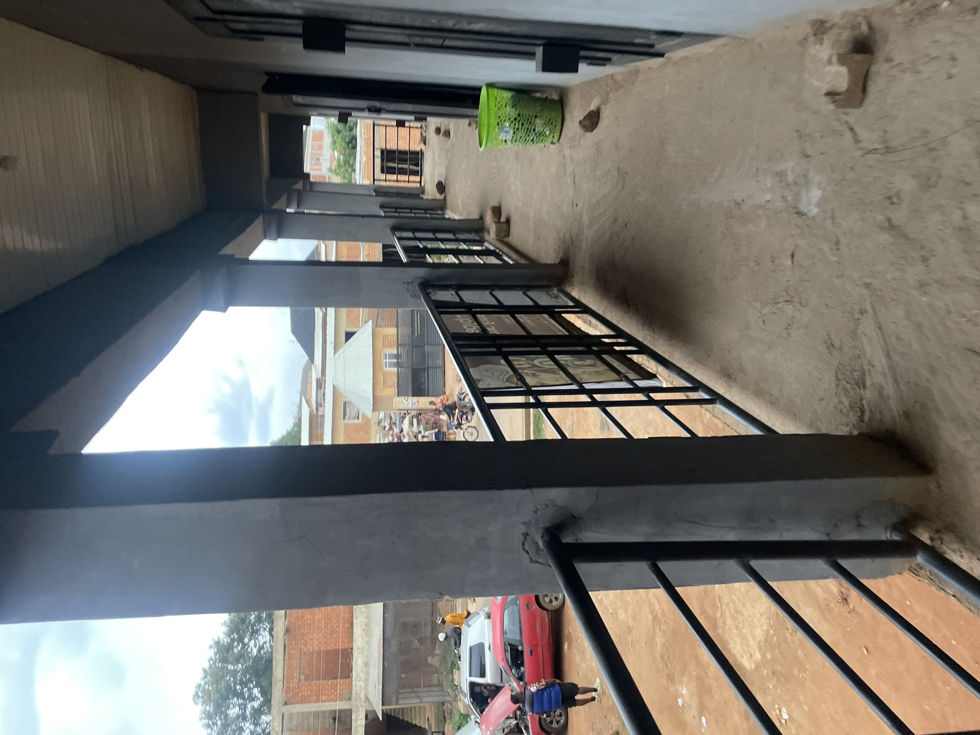 The image depicts a ground-level exterior view of a building in Bukuru, Nigeria, likely a commercial or residential structure. The foreground is dominated by a concrete walkway with a black metal railing. This walkway extends into the distance along the side of the building, which features a series of dark doorways. A bright green plastic trash can is visible on the walkway near the railing, along with several bricks scattered on the ground.
Beyond the railing, an open area with reddish-brown dirt is visible. Several vehicles are present in this area, including a red car with its hood open and a silver SUV. People are seen around these vehicles, with one person wearing a blue vest and shorts standing near the red car, and another individual in an orange shirt and hat near the silver SUV. Further in the background, more buildings constructed with brick and metal sheeting are visible, suggesting an urban or semi-urban environment. Some trees are also present in the distance. The sky is overcast with clouds. The overall scene suggests a possibly unfinished or utilitarian construction area within the city.