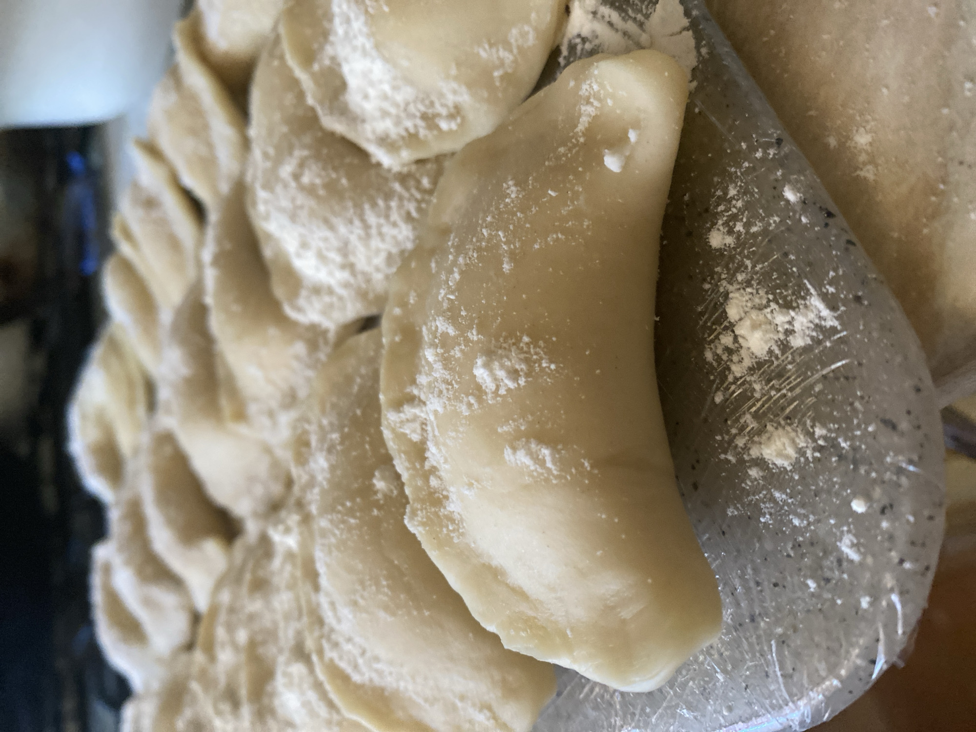 A close-up, eye-level shot shows a tray of uncooked pierogi. The pierogi are arranged in rows, with some covered in a light dusting of flour. The pierogi are crescent-shaped and appear to be made of a pale dough. The background is blurred, but appears to be a kitchen setting with a dark countertop and a white pitcher in the distance. The overall impression is one of homemade food preparation.