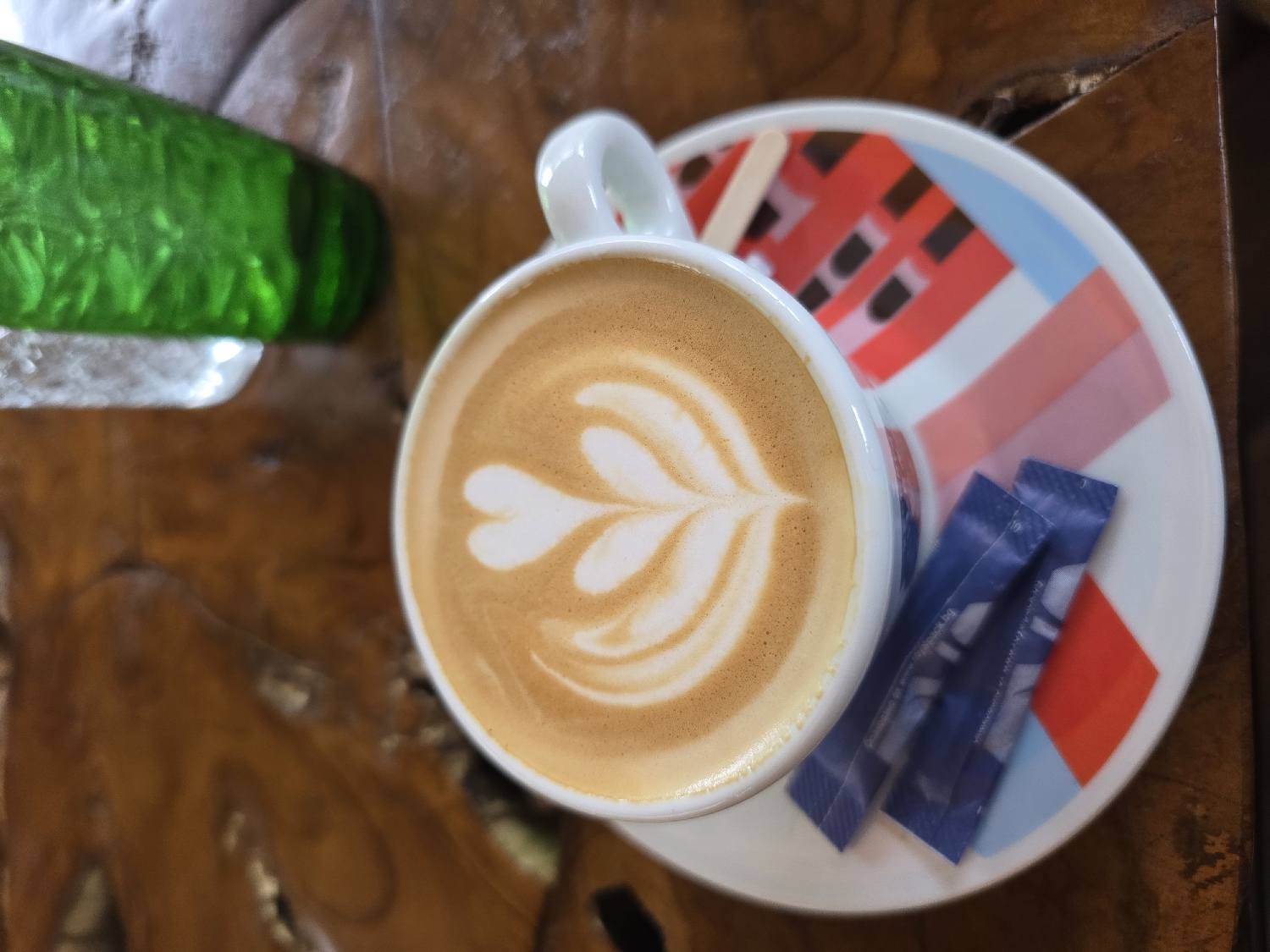 This is an overhead shot of a latte served on a wooden table, likely in a cafe in Burgas, Bulgaria. The main subject is a white ceramic cup holding a latte with intricate white latte art, resembling a heart or a stylized leaf, against the light brown foam.
The cup is placed on a distinctive white ceramic saucer adorned with a modern, abstract geometric pattern featuring blocks and lines in red, pink, and light blue. On the saucer, beside the cup, are two small, dark blue foil packets, presumably sugar, with faint, illegible white text, and a thin wooden stirring stick.
In the upper-left corner of the frame, a vibrant green glass object, possibly a bottle or vase, is partially visible, adding a bright color accent. The entire setup rests on a rich, warm-toned wooden table, showing visible grain and natural textures, which provides a rustic yet cozy backdrop. The lighting is soft and even, suggesting an indoor setting, creating a peaceful and inviting atmosphere. No people are visible, emphasizing the still life arrangement of the coffee service.