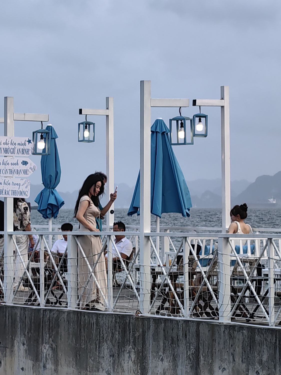 The image depicts an outdoor leisure area, likely a cafe or restaurant situated on an elevated platform or pier, overlooking a body of water in Bai Chay Ward, Vietnam. The sky is overcast, suggesting a cloudy day or late afternoon, with artificial lights already illuminated.
In the foreground, a woman with long, dark hair, dressed in a long, light-colored (beige/cream) dress, stands by the railing, holding a smartphone, possibly capturing a selfie or video. To her right, another woman with her hair in a bun is seated at a table, wearing a light-colored top and looking towards the camera's general direction. Several other patrons, including men in white shirts and a person in a floral-patterned shirt, are seated at tables in the background.
The platform features a white wooden structure with criss-crossed railings and vertical posts. From these posts hang decorative square lantern-style lights with illuminated bulbs. Several closed, blue umbrellas (parasols) are also attached to the posts, suggesting shade provisions. Below the platform, a textured concrete wall forms the base.
The background reveals a calm sea or bay, with distant, misty landmasses that appear to be karst formations, typical of the region around Ha Long Bay.
Visible Vietnamese text on a white sign includes phrases such as "Y NHỚ GIỮ AN BÌNH!" (Remember to keep peaceful!), "biển xanh" (blue sea), "Y CÂN THẬN BẠN NHÉ!" (Be careful, friend!), and a partial message related to waste disposal, "DE RÁC THEO DO". The overall mood appears relaxed and casual.