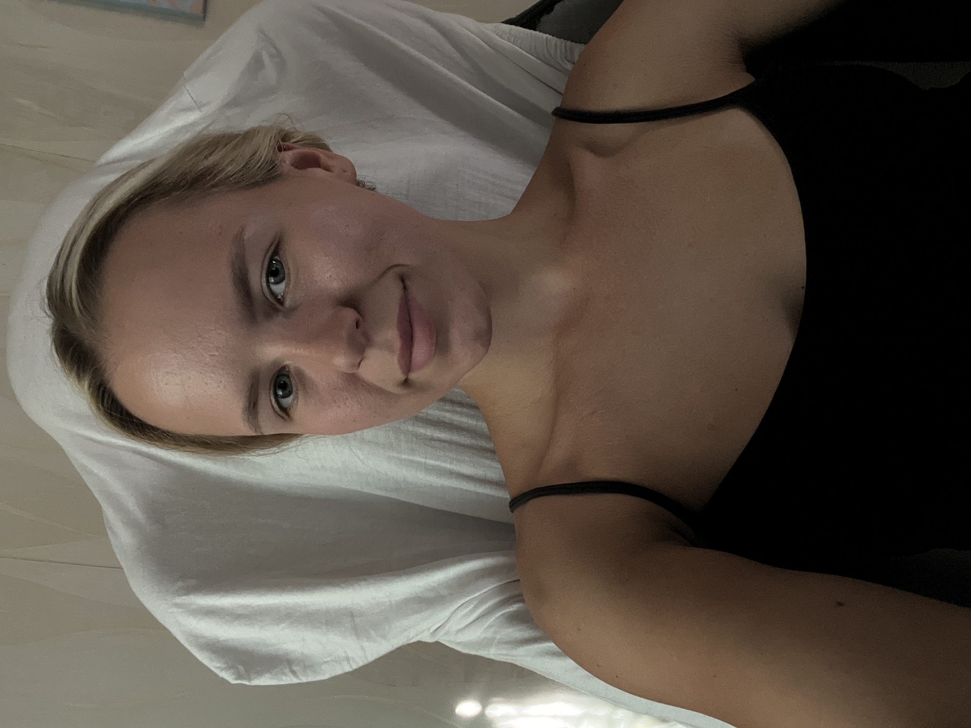An image shows a close-up of a young blonde woman's face and upper body. She is looking directly at the camera with a slight smile. She has blue eyes and fair skin with some redness on her cheeks. She is wearing a black tank top. Behind her, a white fabric is draped, creating a soft, diffused background with some shadows and highlights. The lighting suggests an indoor setting, possibly a bedroom or a studio. There are no visible indications of the city or specific location.