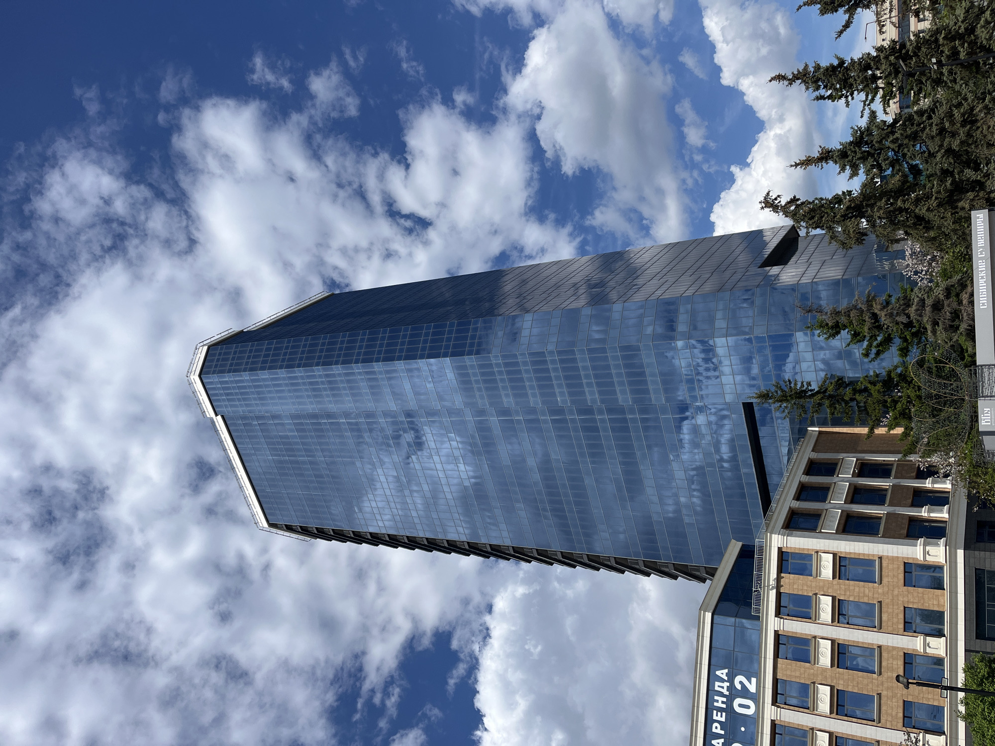 The image shows a tall, glass-clad skyscraper in Krasnoyarsk, Russia, under a partly cloudy blue sky. The building's facade reflects the clouds. Adjacent to the skyscraper is a smaller, multi-story building with a brown and beige facade and blue windows. The word "АРЕНДА" is vertically printed on its glass entrance. Green trees are partially visible on the right side of the image, and a sign with Cyrillic text, likely advertising the "Sibirskie Suveniry" (Siberian Souvenirs) store, is also present. The angle is from a low perspective, looking up at the buildings and sky.