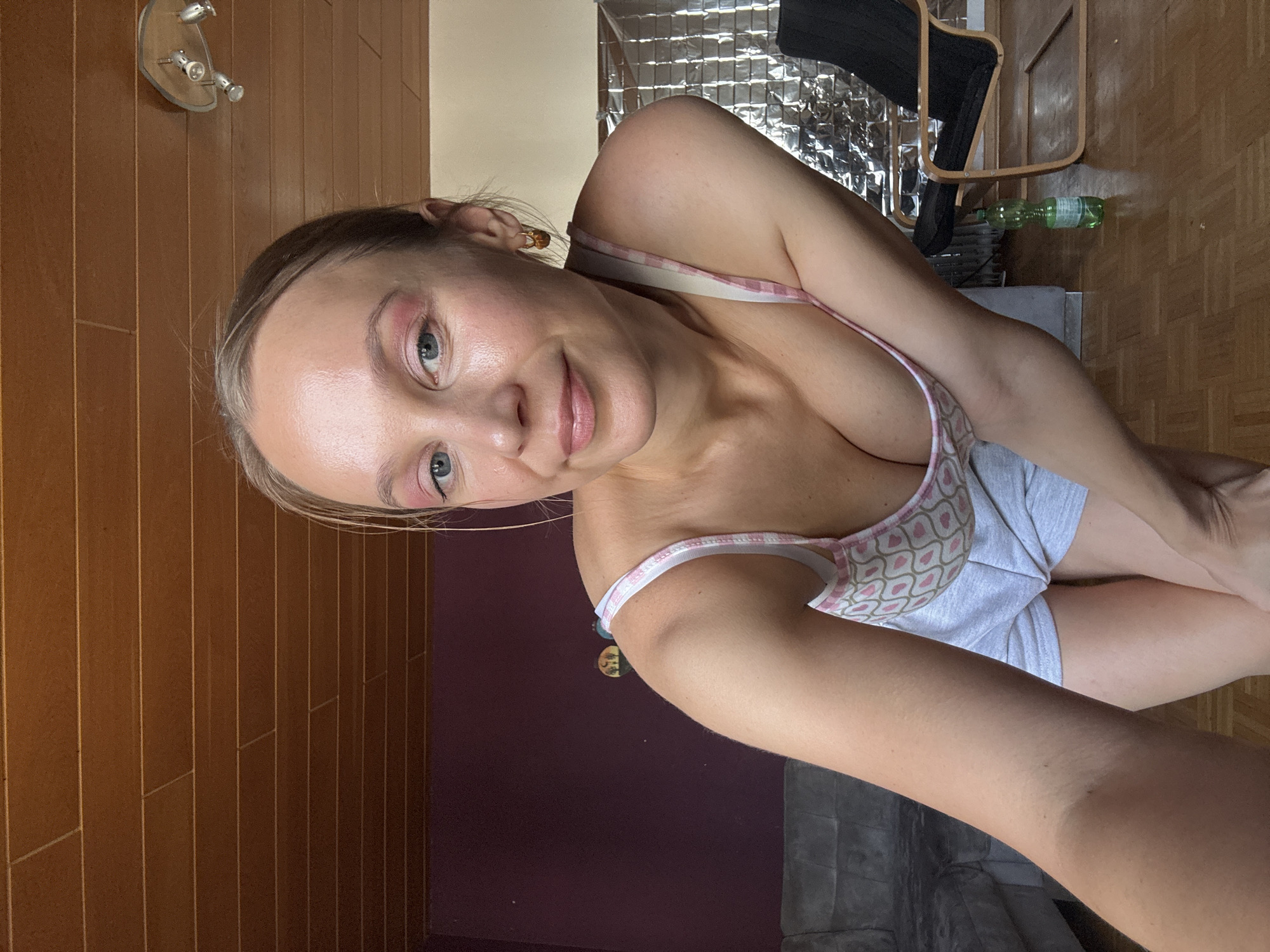 This media file features a young woman with blonde hair, posing for a selfie. She is wearing a light-colored top with a pink and white checkered strap and a patterned design of hearts. Her makeup includes pink eyeshadow. The background suggests an indoor setting, possibly a living room or studio, with wooden flooring and walls. Visible objects include a chair with a reflective silver material behind it, a green plastic bottle, and a light fixture mounted on the wall. The lighting appears to be artificial, suggesting it could be daytime or evening. The woman's expression is a slight smile, and she is looking towards the camera. There is no visible text in the image. The context indicates the location is Überherrn, Germany.