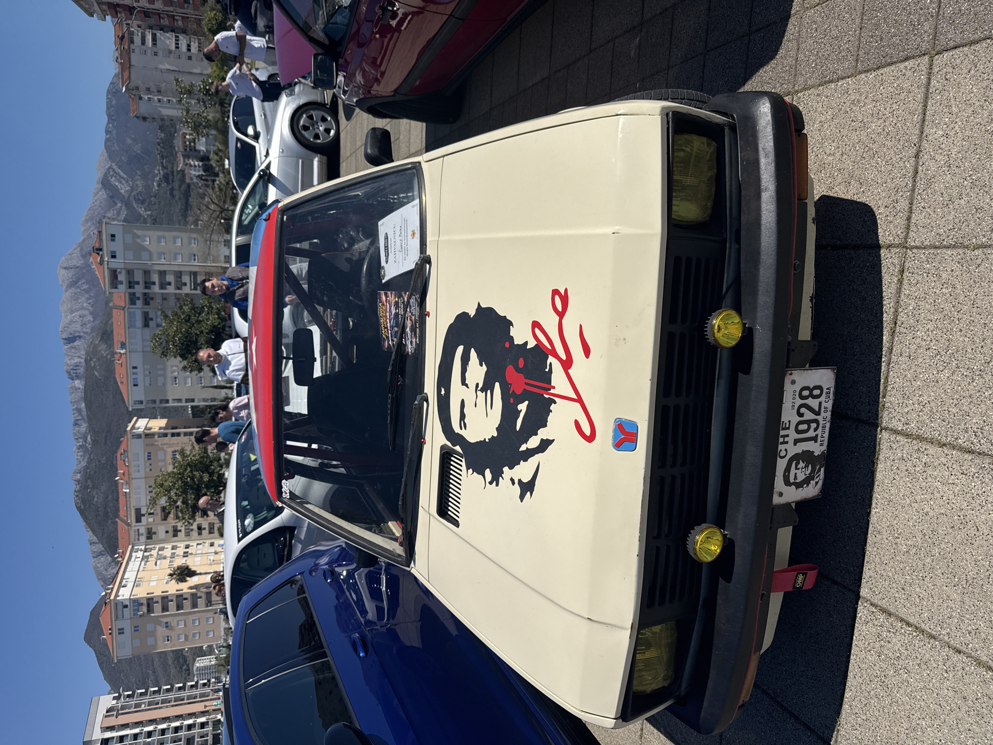 A slightly rotated, eye-level, full outdoor shot depicts the front of a cream-colored car adorned with a black and red Che Guevara decal on the hood, parked on concrete paving stones. The car's front grill is black with yellow fog lights, and it bears a license plate reading "CHE 1928 REPUBLIC OF CUBA."
Behind the cream-colored car is a blue car, and beyond that are glimpses of other vehicles and people. In the background, several multi-story apartment buildings are visible, set against a mountainous landscape under a clear blue sky. The photograph suggests a sunny day in an urban environment, specifically identified as the city of Bar, Montenegro, given the context. Several individuals can be seen standing near the cars, although their actions and interactions are not clearly discernible.