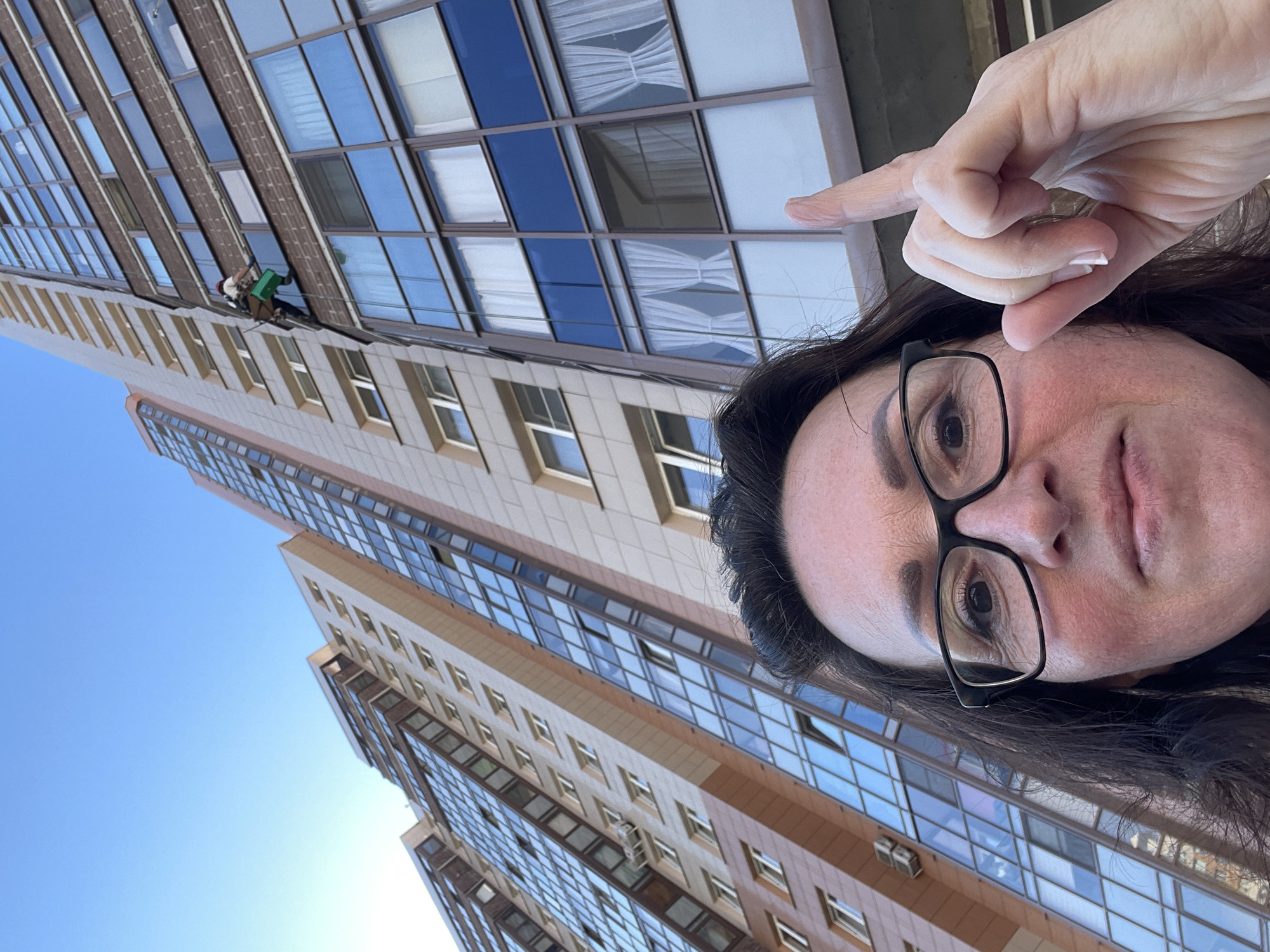 The image presents a low-angle, upward-facing perspective of a modern urban environment in Saint Petersburg, Russia. In the immediate foreground, a woman with dark hair and black-framed eyeglasses is captured from her forehead to her chin. Her right eye is visible, and her skin exhibits light tones with some reddish coloration on her cheeks. Her right hand is raised beside her head, with the index finger extended directly upwards, pointing towards the buildings above.
Dominating the mid-ground and background are two tall, multi-story residential or office buildings, characteristic of modern urban architecture in Saint Petersburg. The primary building on the right features a facade composed of alternating sections of light beige and dark brown brickwork, interspersed with numerous large, rectangular glass windows. Many of these windows reflect the clear blue sky, while others reveal white internal curtains.
Approximately several stories up on the main building, a window cleaner is visibly suspended by a system of ropes and harnesses. The cleaner is equipped with a light-colored hard hat featuring a red band, and is seen holding or positioned near a green bucket or cleaning apparatus. This individual is actively working on a window pane set within a darker brown section of the building facade.
To the left, a second, similarly tall building is partially visible, sharing architectural elements such as numerous windows and a beige-toned facade. The sky above the buildings is uniformly clear and bright blue, indicating fair weather conditions. The scene captures a moment of urban activity, with a citizen observing and highlighting the work of a high-rise window cleaner.