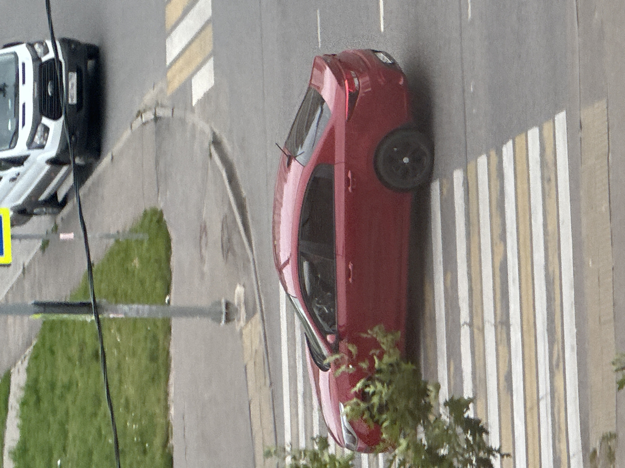 A red sedan is crossing a pedestrian crosswalk on a street in Podolsk, Russia. The car is a Kia Rio, based on its distinctive taillights and body shape. The car is traveling from left to right in the frame and is partially obscured by a green bush in the foreground. In the background, a white Ford Transit van is stopped at a traffic light. The street is paved with asphalt, and there is a sidewalk to the left of the street with grass and a lamppost. The sky is overcast, and the lighting suggests it is daytime.