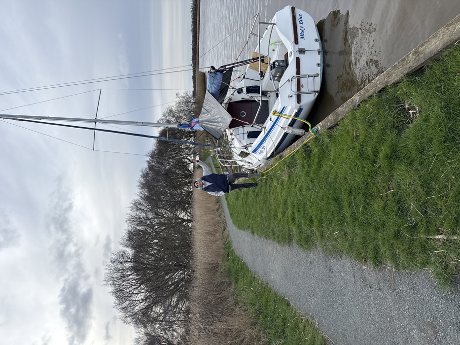 An outdoor scene captured on an overcast day depicts a male individual standing beside a moored white sailing yacht on a calm waterway, likely located within the Norfolk Broads in the district of North Norfolk, United Kingdom. The man, with dark hair and a beard, is positioned on a narrow strip of green grass separating a gravel path from the water. He wears a dark sleeveless puffer vest over a light-colored long-sleeved shirt and dark trousers, and is facing the camera with a subtle smile, his right hand slightly raised in a gesture. The sailboat, displaying the name "Misty Blue" on its transom and "Regal 525" on its hull, features a single mast, an outboard motor mounted on the stern, and a grey tarp covering a portion of its cabin area. Yellow mooring lines connect the boat to the wooden embankment. A light grey gravel path curves gently to the left alongside the grassy bank. The background consists of tall, dry, light-brown reeds and a dense line of leafless deciduous trees. Distant reed beds and a faint structural silhouette are visible on the far right horizon under a cloudy sky. The water is brownish and still.