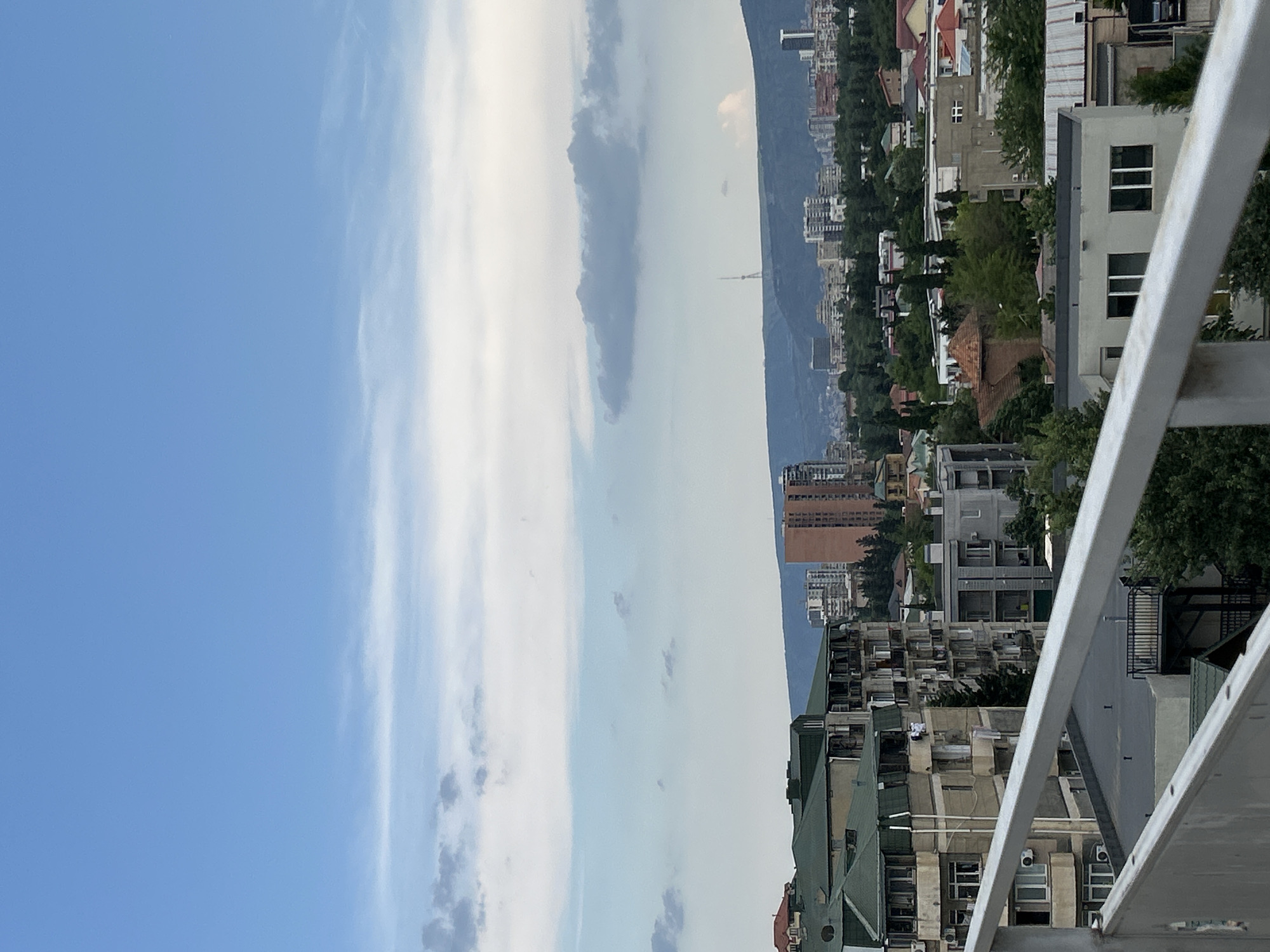 The image depicts a panoramic view of a city nestled against a backdrop of distant mountains. The foreground is dominated by a metal railing, suggesting the vantage point is from a balcony or elevated structure. Below the railing, a dense urban landscape unfolds, characterized by a variety of buildings, ranging from multi-story apartments with visible windows and air conditioning units to smaller, older structures with pitched roofs. Lush green trees are interspersed throughout the city, softening the urban sprawl.
In the mid-ground, a tall tower with a distinctive antenna structure rises above the cityscape, silhouetted against the hazy sky. The city extends towards the base of a large mountain range, which appears somewhat obscured by clouds. The sky itself displays a mix of bright blue and wispy white clouds, suggesting fair weather with some cloud cover. The lighting indicates it is likely daytime, possibly late morning or afternoon, given the brightness and the shadows.
The scene conveys a sense of urban life viewed from a distance. There are no visible people in the frame. The overall impression is one of a sprawling city coexisting with natural mountainous terrain, under a bright, somewhat cloudy sky. The angle of the shot, with the railing prominently in the foreground and the city stretching horizontally, emphasizes the scale of the urban environment.