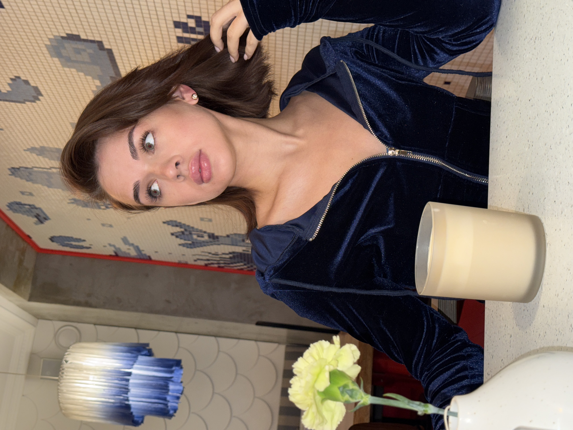 A young woman with shoulder-length, dark brown hair and light-colored eyes is captured in an indoor setting in Perm, Russia. She is looking upwards and to her right with a thoughtful or slightly pensive expression, her head tilted. Her left hand is raised, gently touching her hair near her ear. She wears a dark blue, possibly velvet or velour, hooded jacket or top with a silver zipper that is partially unzipped. A small, round, light-colored stud earring is visible in her left ear.
To her right, on a light-colored table, sits a frosted glass candle holder containing a light-colored candle. Further to the right, a single light yellow carnation flower with a green stem is arranged in a small, white ceramic vase.
The background features a distinctive patterned ceiling or wall, composed of small tiles forming geometric shapes and abstract designs in shades of beige, cream, and various blues and grays. A red border runs along the edge of this patterned surface. Below it, a darker, solid section gives way to a lighter wall with a subtle, repeating scalloped or circular relief pattern, which also shows a light fixture with blue and white stripes. The overall lighting is soft and warm, suggesting an evening ambiance in what appears to be a cafe or restaurant. No visible text is present in the image.
