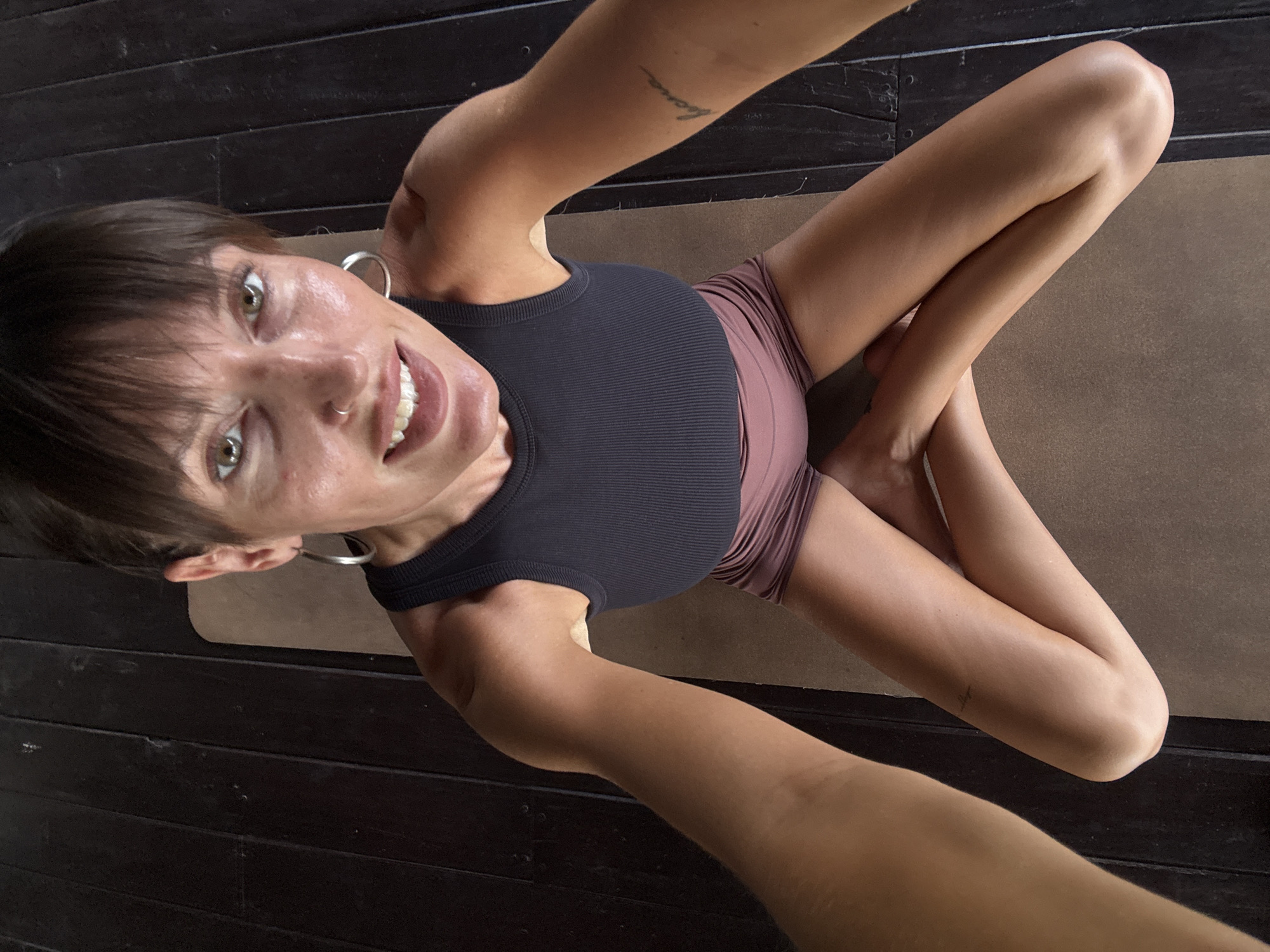 A high-angle selfie shows a fair-skinned woman seated cross-legged on a light brown yoga mat, which lies on dark brown, horizontally-laid wooden floorboards. The woman wears a black ribbed tank top and pinkish-brown shorts. She has short dark hair with bangs, hazel eyes, and a nose ring. She is smiling, showing her teeth, and wearing large silver hoop earrings. Tattoos are visible on her left arm. The image was taken in Mengwi, Indonesia.