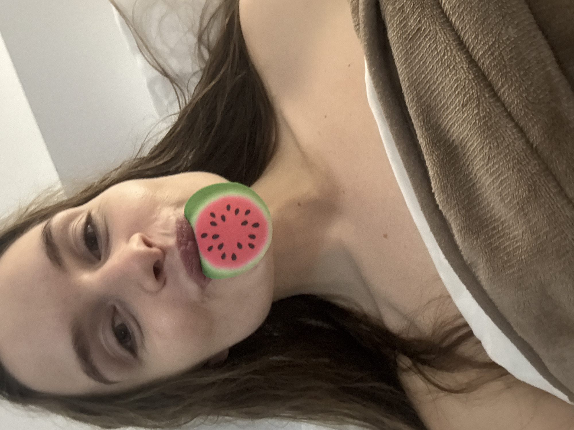 A woman with brown hair is lying on her side, partially covered by a brown blanket with a white edge. A watermelon-shaped object is placed near her mouth. The location is not identifiable from the image.