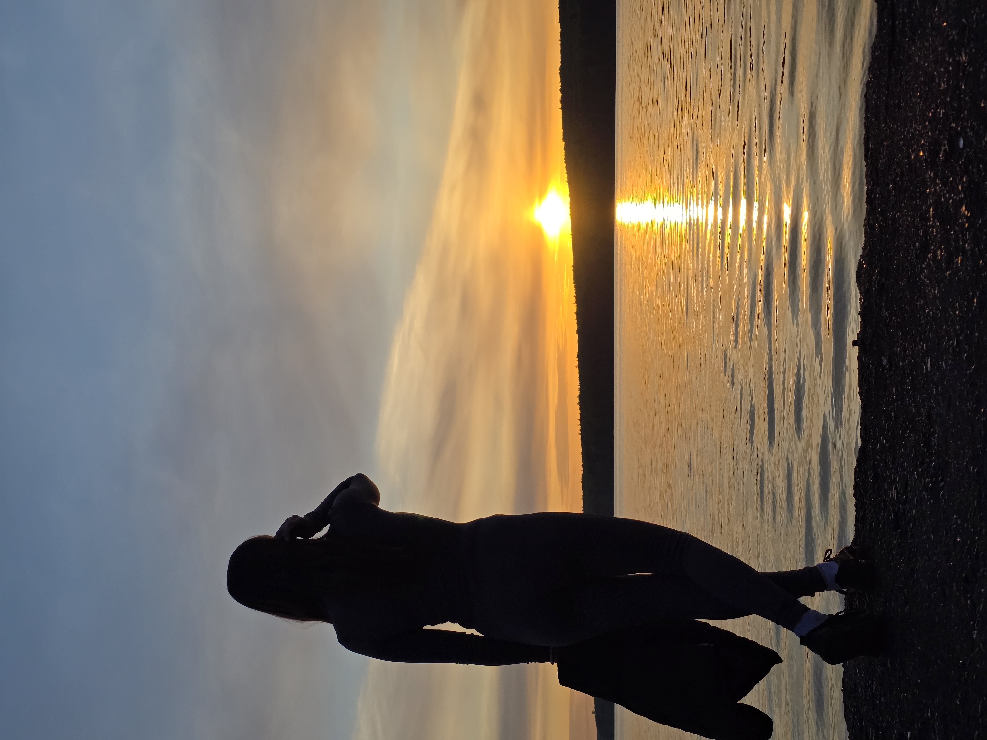 The image depicts a silhouette of a person standing near a body of water at sunset. The location is likely Chaikovsky, Russia based on the presence of the Kama River and the typical Russian climate. The person, seen in silhouette, is slender and wears long sleeves and pants, possibly leggings. They have long hair and are standing with their arms near their head, perhaps adjusting their hair or taking a photo. They are standing on a rocky shore next to a large body of water. The sun is setting in the distance, creating a golden glow on the water and the sky. The sky is filled with light clouds. There is a tree line on the opposite side of the water. The sun's reflection creates a distinct line on the water surface. The scene is peaceful and serene.