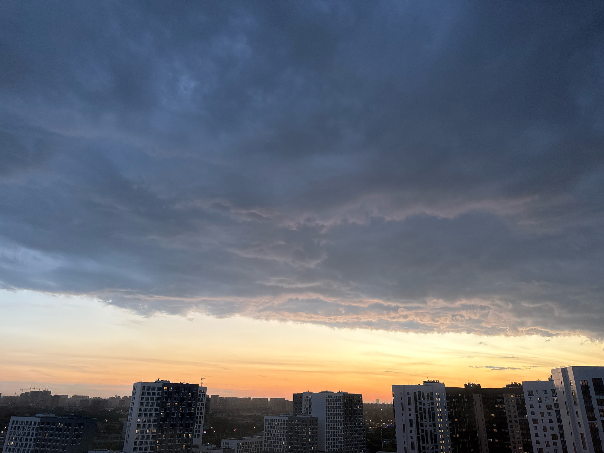 The image depicts a cityscape at dusk, likely in Lyubertsy, Russia. Dark, heavy clouds dominate the upper half of the frame, with a band of lighter, pinkish-orange clouds near the horizon. Below the clouds, the sky transitions to a warm orange and yellow gradient. Silhouetted against the sky are numerous multi-story buildings, characteristic of a dense urban environment. Several tall, modern apartment buildings with visible windows are prominent in the foreground and mid-ground, some with dark facades and others with lighter exteriors. In the distance, a hazy urban sprawl is visible, interspersed with construction cranes. The overall scene conveys a sense of impending weather or the end of a day in an urban setting. There are no discernible people or specific actions taking place.