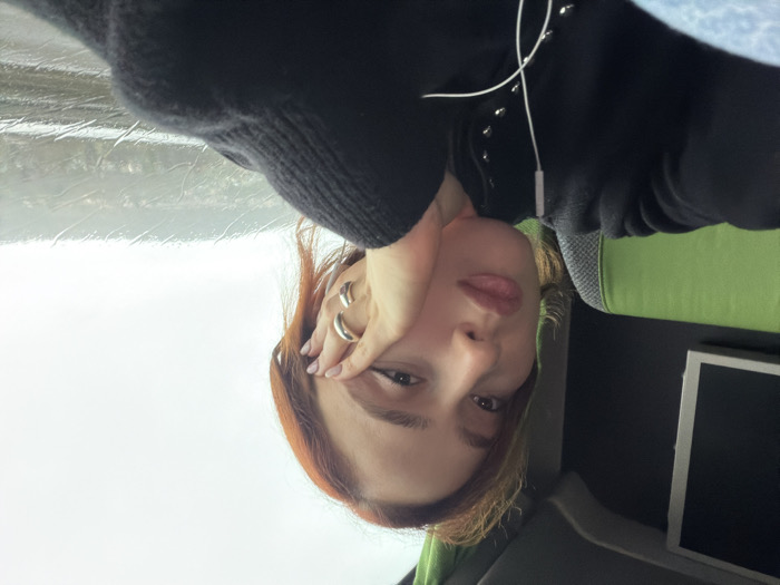 A female subject with reddish-brown hair is depicted in a medium-shot, seated inside a vehicle. The location is stated as Moineville, France, though no specific geographical identifiers are visible in the image. The subject is looking directly forward. Her right hand, adorned with two silver-colored rings on the ring and middle fingers, is positioned against her cheek. She wears a dark, long-sleeved top featuring multiple metallic studs down the front. White wired earbuds are present, with one earpiece in her left ear. To her left, a portion of a green and grey upholstered seat is visible, along with a dark wall housing a white-framed rectangular screen. The right side of the frame features a window displaying an overcast exterior. Rain or condensation streaks are visible on the windowpane, partially obscuring a distant, hilly landscape under a bright, diffused sky. The interior lighting is subdued.