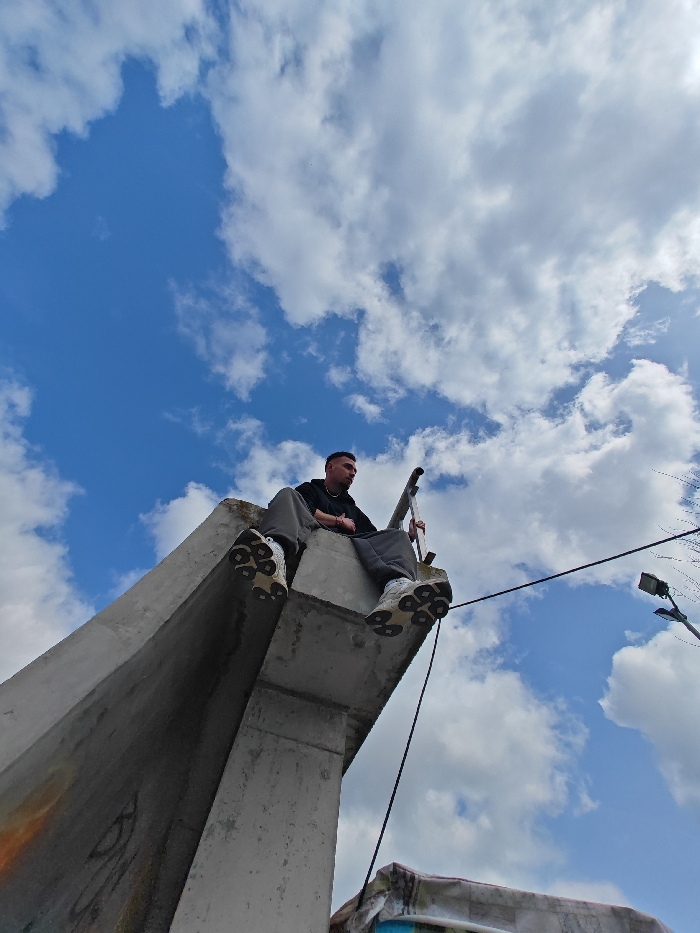 This image, taken in Shumen, Bulgaria, presents a low-angle perspective of a lone male subject seated on a massive, weathered concrete structure. The male, facing right (viewer's left), is dressed in a dark hooded sweatshirt, grey sweatpants, and white athletic shoes with pronounced, textured soles. His right hand grips a vertical metal rail or ladder adjacent to him, while his legs dangle off the edge of the concrete beam. The concrete structure exhibits a brutalist aesthetic with visible weathering and minor discoloration. A black cable descends from the structure in front of the subject's feet. The background consists of a bright blue sky with scattered white cumulus clouds. To the lower right, a modern street light fixture on a pole is visible, along with the upper edge of another concrete element displaying light blue graffiti.