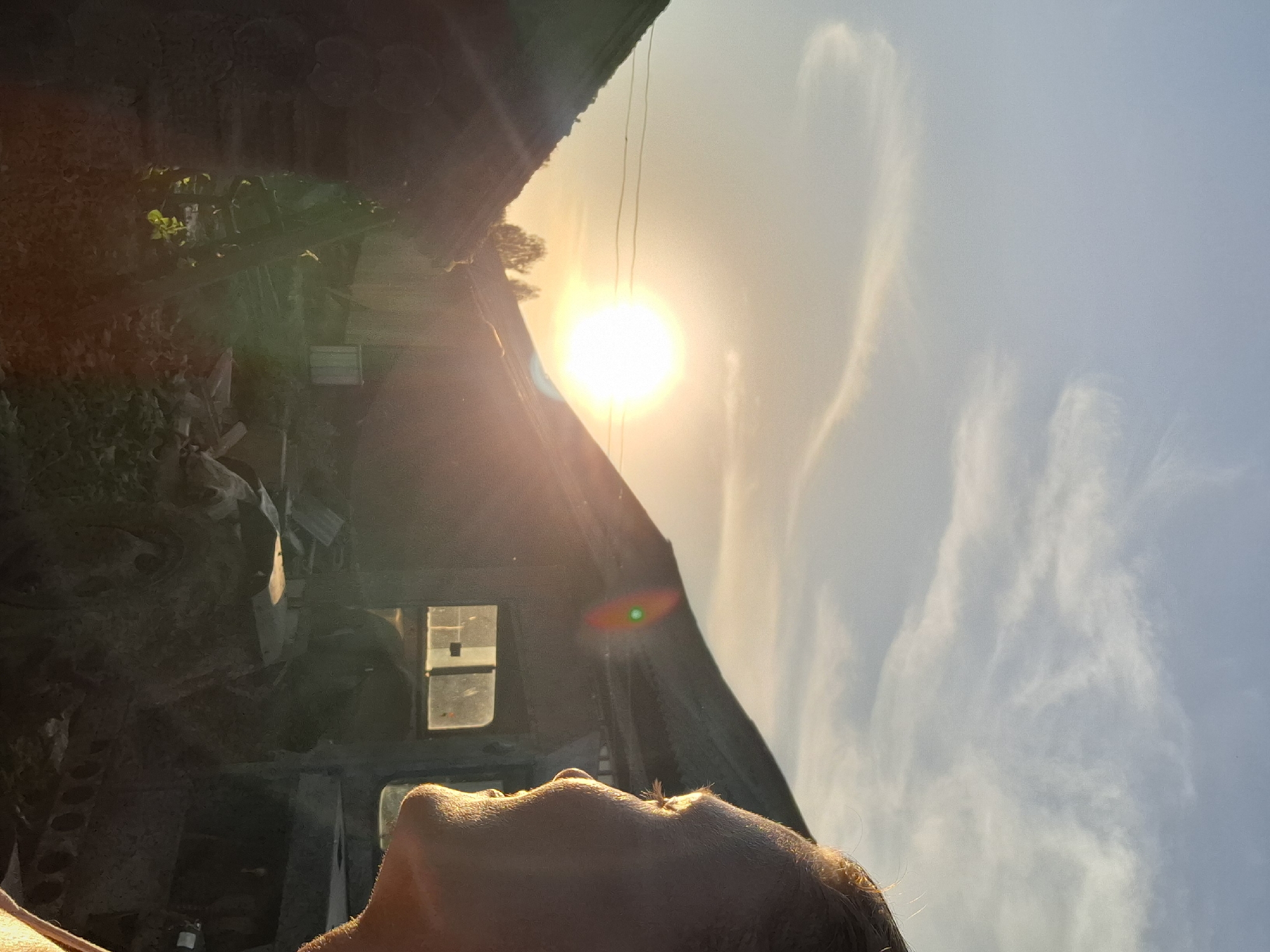 The image shows a low-angle view, looking up. In the foreground is the partial view of a person's face. The person is looking toward a bright sun that is partially obscured by a structure resembling a building. There are also some white clouds in the sky. The area around the building looks like a cluttered area. Based on the specified location, this appears to be a snapshot in or near Шешурга, Russia.