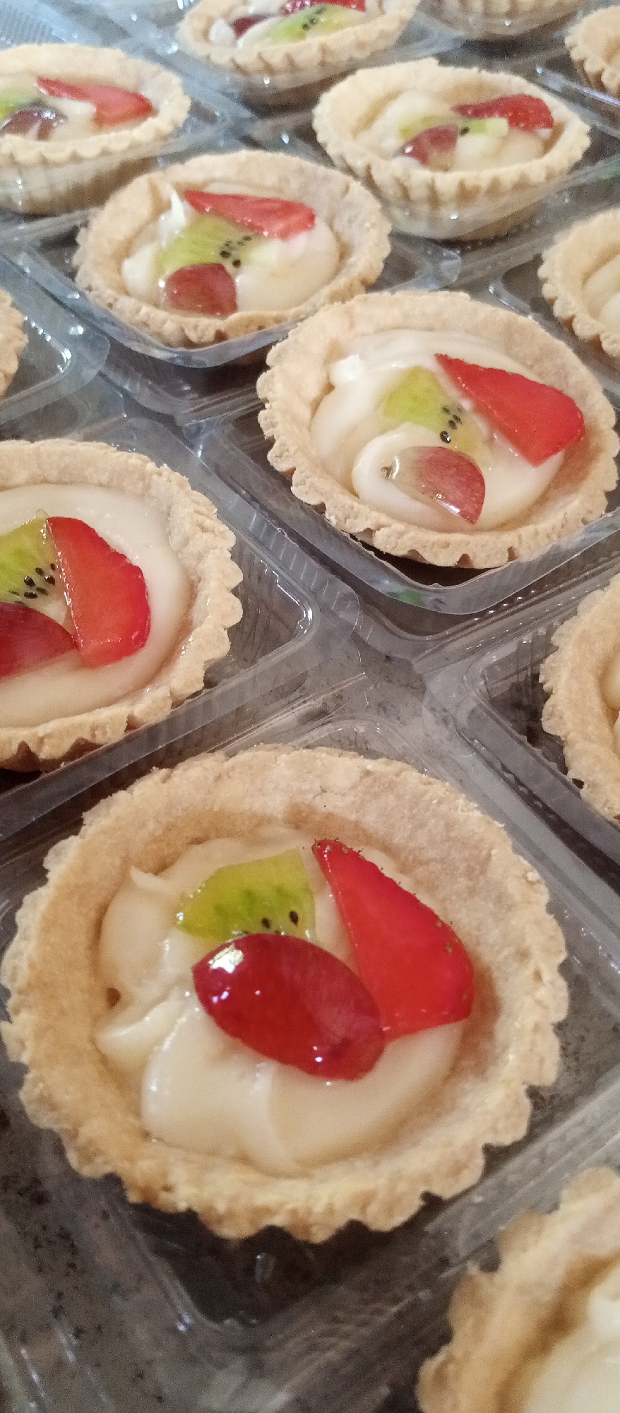 The image displays a close-up, overhead view of multiple individual fruit tarts arranged in clear plastic packaging. The tarts have a light brown, crimped crust filled with a creamy white custard. Toppings on the custard include slices of red strawberry, green kiwi with visible seeds, and pieces of red grape. The tarts are organized in a grid-like fashion within the transparent containers. The background is blurred and appears to be a dark, textured surface. The lighting is diffused, highlighting the textures and colors of the fruit and crust. There are no people visible in the image. The setting is likely a food display or preparation area.