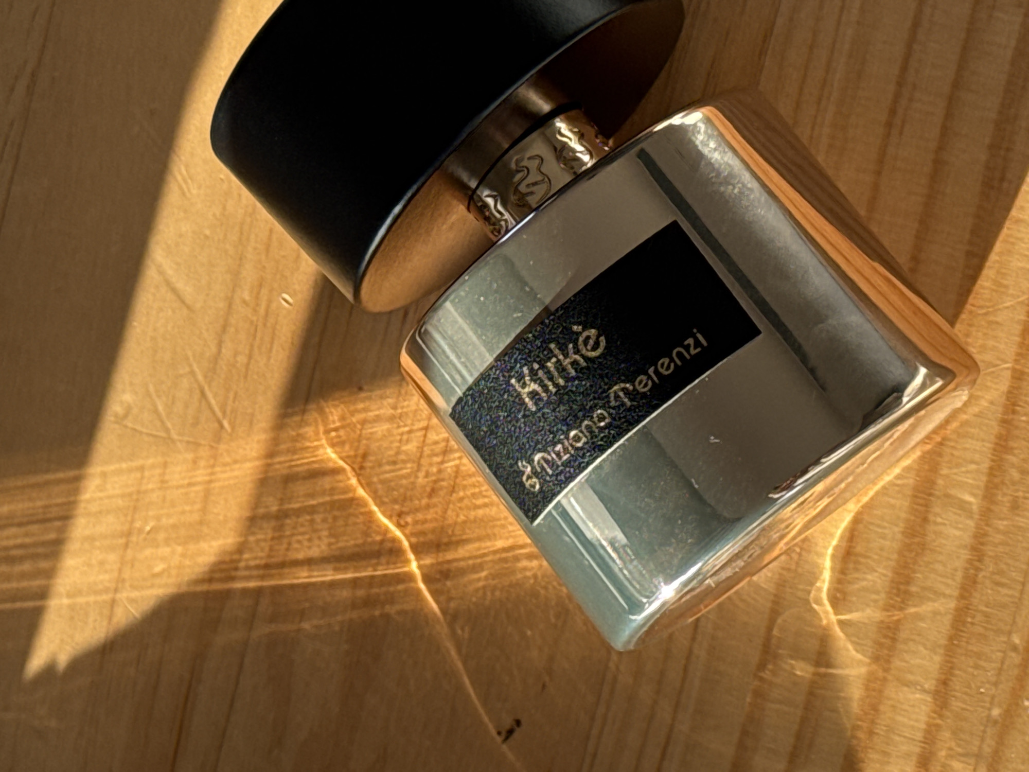 A close-up, high-angle shot shows a silver perfume bottle with a black label and cap lying on a wooden surface. The bottle is angled, with the cap to the upper right. The black label on the bottle has text in a serif font, possibly featuring the words "Kirke" and "Tiziana Terenzi". The wooden surface has visible grain and is illuminated by diagonal shafts of sunlight, creating bright reflections and shadows. The overall impression is of a luxurious product displayed in natural light. The location is not specified in the image.
