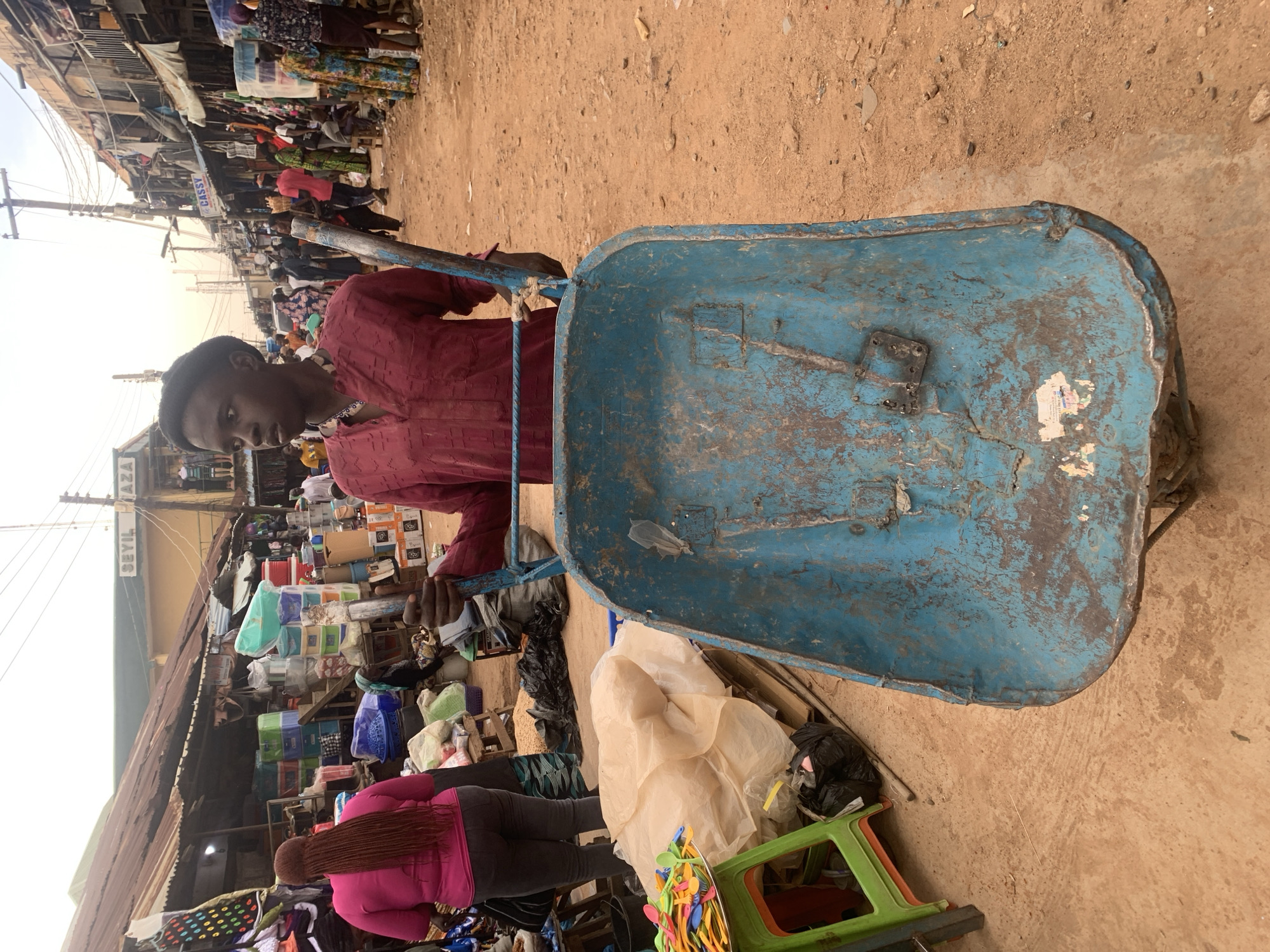 This image captures a vibrant, bustling street market scene, characteristic of an open-air market in Jos, Nigeria.
The central subject is a young man, likely in his late teens or early twenties, positioned slightly left of center. He wears a maroon or dark red long-sleeved garment with a textured pattern and a dark cap. He holds the handles of a large, well-worn blue wheelbarrow that dominates the right side of the foreground. The wheelbarrow is visibly aged, with chipped paint, rust, and some attached labels or stickers.
The background reveals a lively market street with numerous people and makeshift stalls. Other individuals are seen browsing or working; notable among them is a woman in a bright pink top with long, braided hair, seen from behind. The market stalls display a variety of goods, including stacks of colorful plastic containers, household items, and what appear to be bundles of clothing or fabric. A small display of colorful plastic spoons is visible in the lower right corner.
The setting is an outdoor market with a dusty, unpaved ground. Utility poles with numerous wires crisscross overhead, and many stalls are covered by simple corrugated iron roofs. The lighting suggests it is daytime, likely a bright morning or afternoon. The overall atmosphere is one of active commerce and daily life.
Visible text includes "SEYIL PLAZA" on a sign in the background to the left, and "CASSY" on another sign further down the street. The young man's expression is calm, almost contemplative, as he stands with his wheelbarrow amidst the market activity.