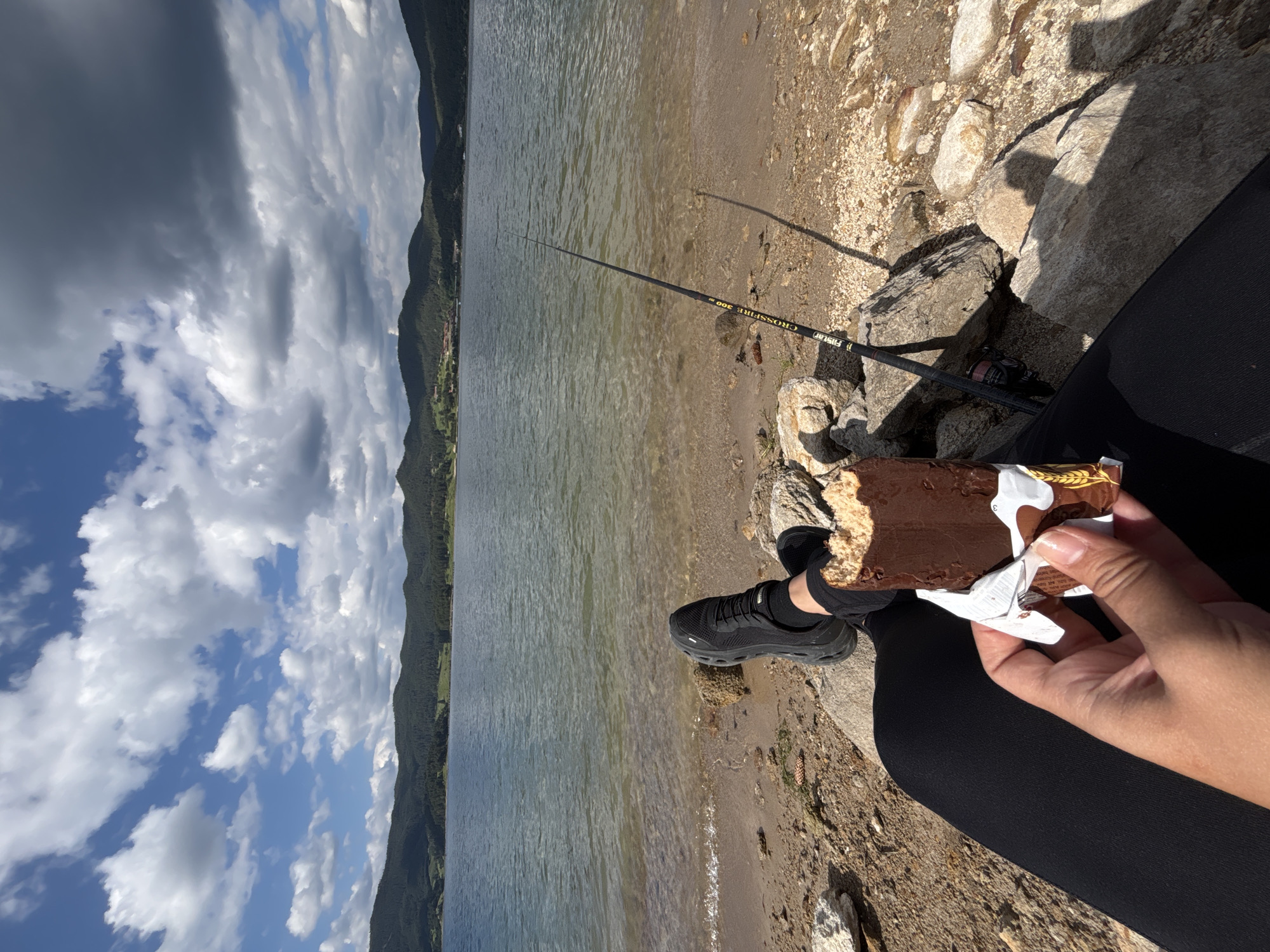 The image shows a person sitting by a lake with a fishing rod. The person is wearing black leggings and black sneakers. They are holding a chocolate-covered ice cream bar. The lake is calm, and the water is clear. Mountains can be seen in the background, covered in trees. The sky is partly cloudy, with dark clouds and patches of blue sky. Sunlight is casting shadows on the ground. The overall scene suggests a relaxed outdoor activity, possibly fishing and enjoying a treat on a warm day. The time of day appears to be afternoon due to the lighting and shadows. The brand name "FiiStar" is visible on the fishing rod, along with the text "CROSSFIRE 300".