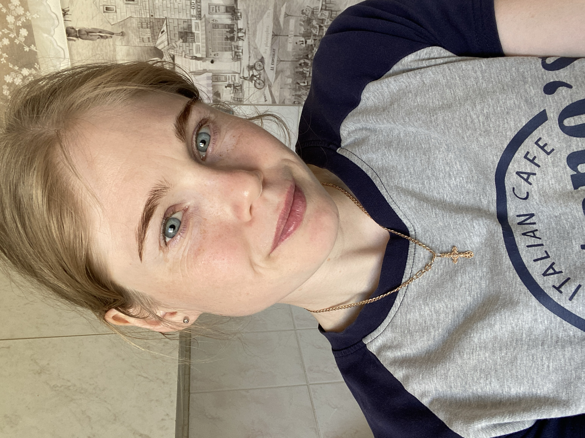 A woman with blonde hair and blue eyes is indoors. She is wearing a grey and navy blue t-shirt, and a gold necklace with a cross pendant. The background includes a tiled wall and a wallpaper with a city scene. The location appears to be within an indoor space, but there is no evidence to suggest she is in the city of Chaikovsky, Russia.