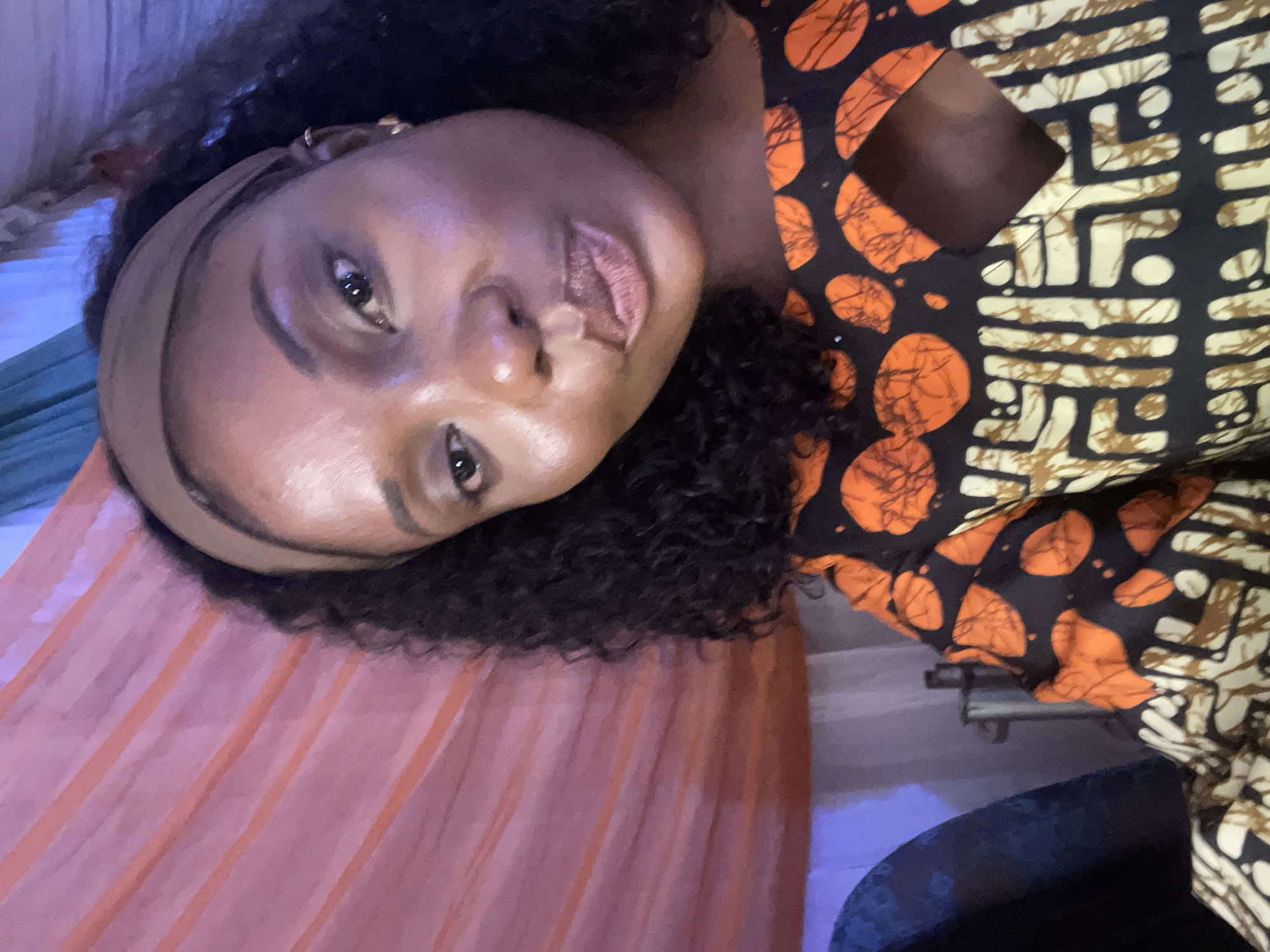 A dark-skinned woman is lying down, tilted towards the camera. She wears a neutral-toned headband and has dark, curly hair. Her clothing features a bold pattern of black, orange, and cream. She appears to be indoors, lying on a surface with orange and pink hues. The background is indistinct but suggests an interior space, potentially in Jos, Nigeria, although there is no definitive evidence to confirm the location. The focus is on the woman's face and clothing.